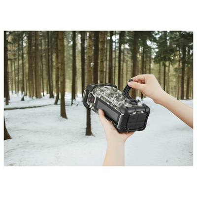 Eine Person hält eine schwarze Wildkamera in einem schneebedeckten Wald und justiert sie. Im Hintergrund sind hohe Bäume zu sehen.