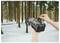 Eine Person hält eine schwarze Wildkamera in einem schneebedeckten Wald und justiert sie. Im Hintergrund sind hohe Bäume zu sehen.
