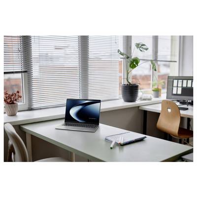 Ein Laptop auf einem Schreibtisch in einem hellen Büroumfeld, mit einem Notizbuch und Stiften in der Nähe, große Fenster und Pflanzen im Hintergrund.