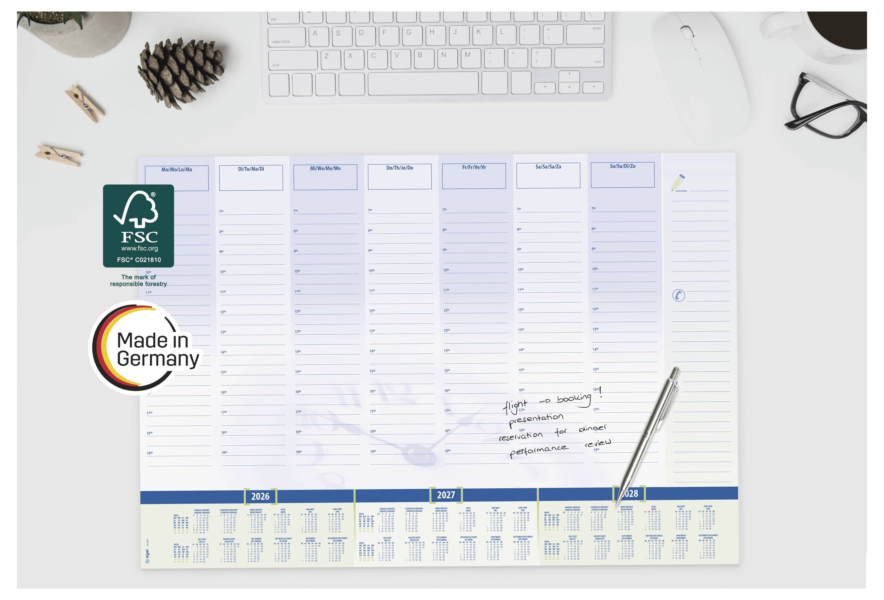 Schreibtischkalenderplaner mit FSC- und „Made in Germany"-Logos, ein Stift, eine Tastatur und Büroartikel auf einem weißen Schreibtisch.