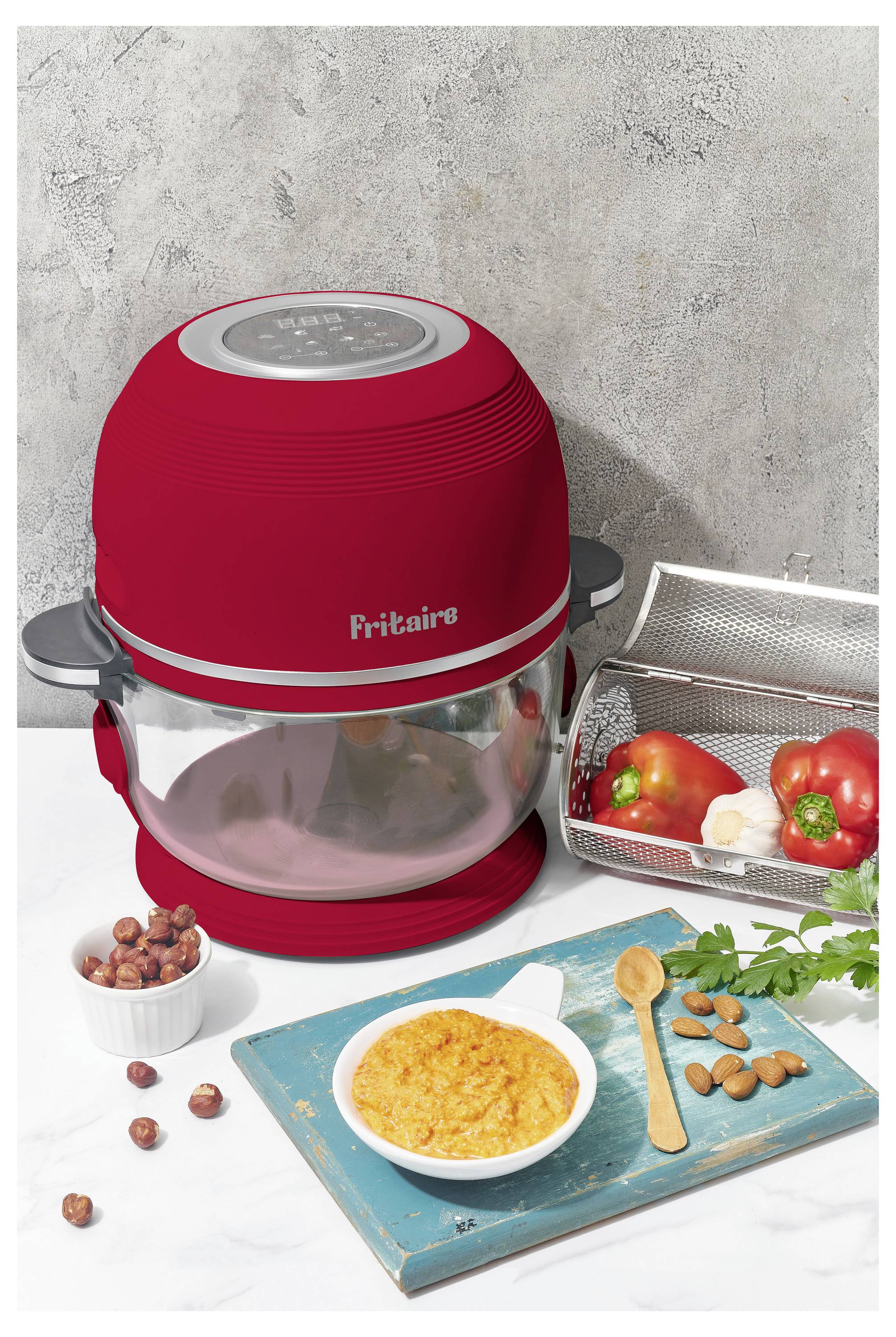Ein roter Heißluftfritteuse mit digitaler Anzeige steht auf einer weißen Arbeitsplatte, daneben frisches Gemüse, Mandeln und eine Schüssel mit Dip auf einem blauen Schneidebrett.