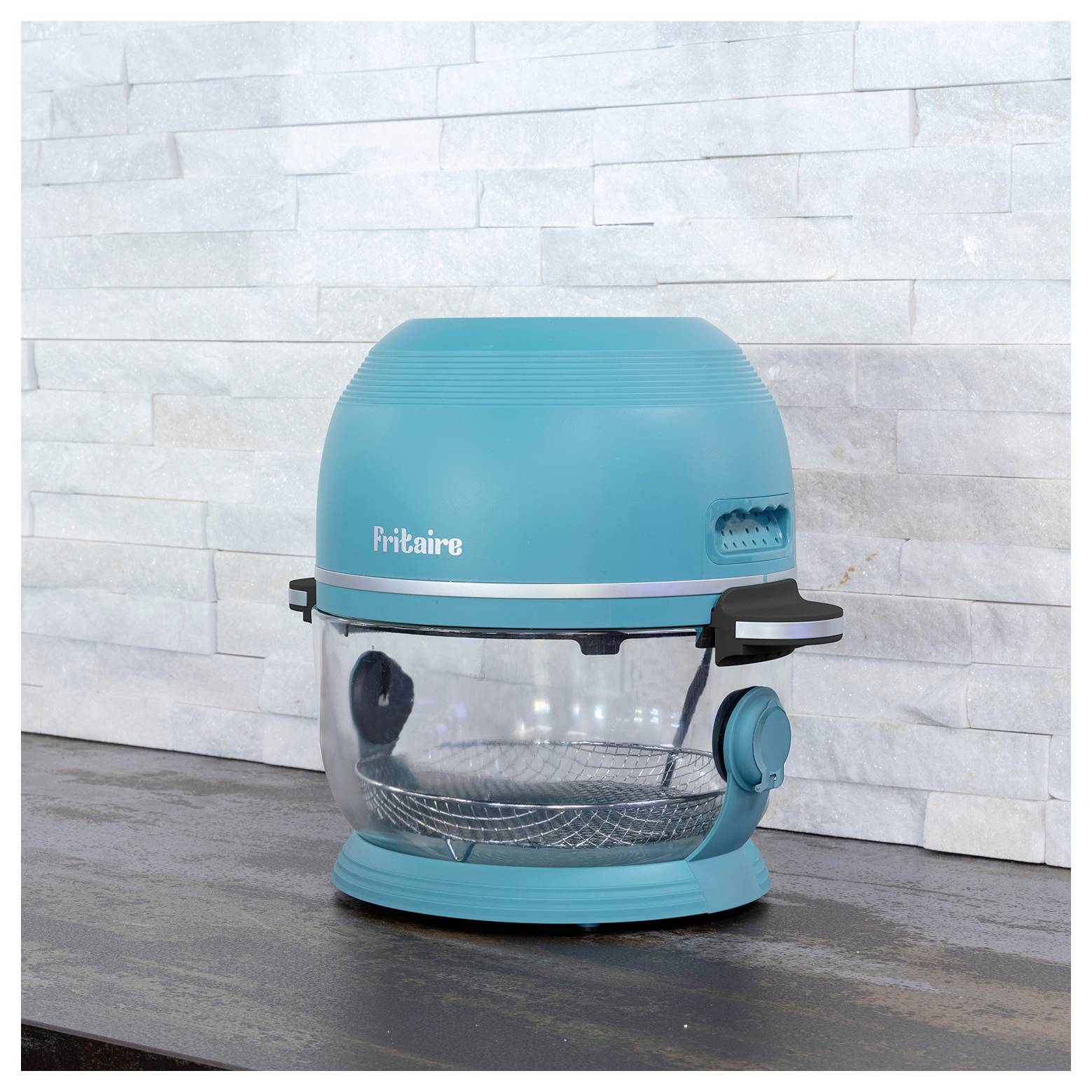 Ein blauer Fritaire-Heißluftfritteuse steht auf einer dunklen Arbeitsplatte vor einem hellen Steinwandhintergrund.