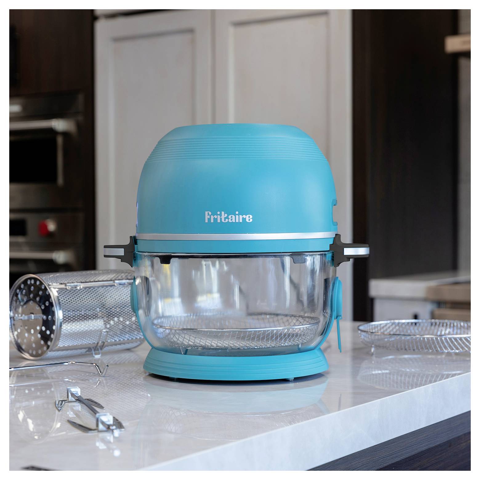 Ein türkisfarbener Heißluftfritteur mit der Aufschrift "Fritaire" steht auf einer Küchenarbeitsplatte. In der Nähe befinden sich Frittierkörbchen und Küchenutensilien, die seine Verwendung beim Kochen unterstreichen.