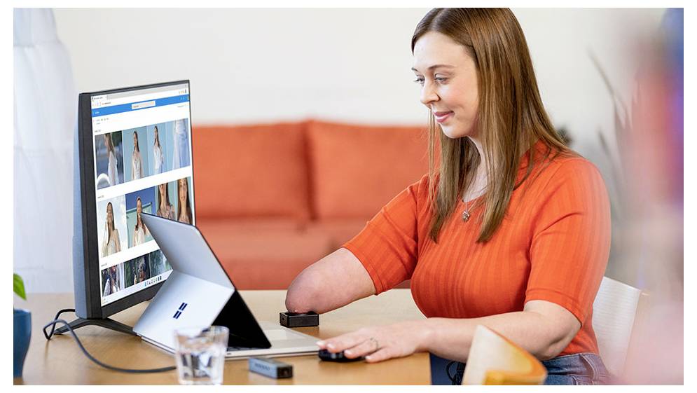Eine Frau nutzt einen Laptop mit einem Hilfsgerät und durchsucht eine Auswahl an Kleidung auf einem großen Monitor, was die Barrierefreiheit in der Technologie demonstriert.
