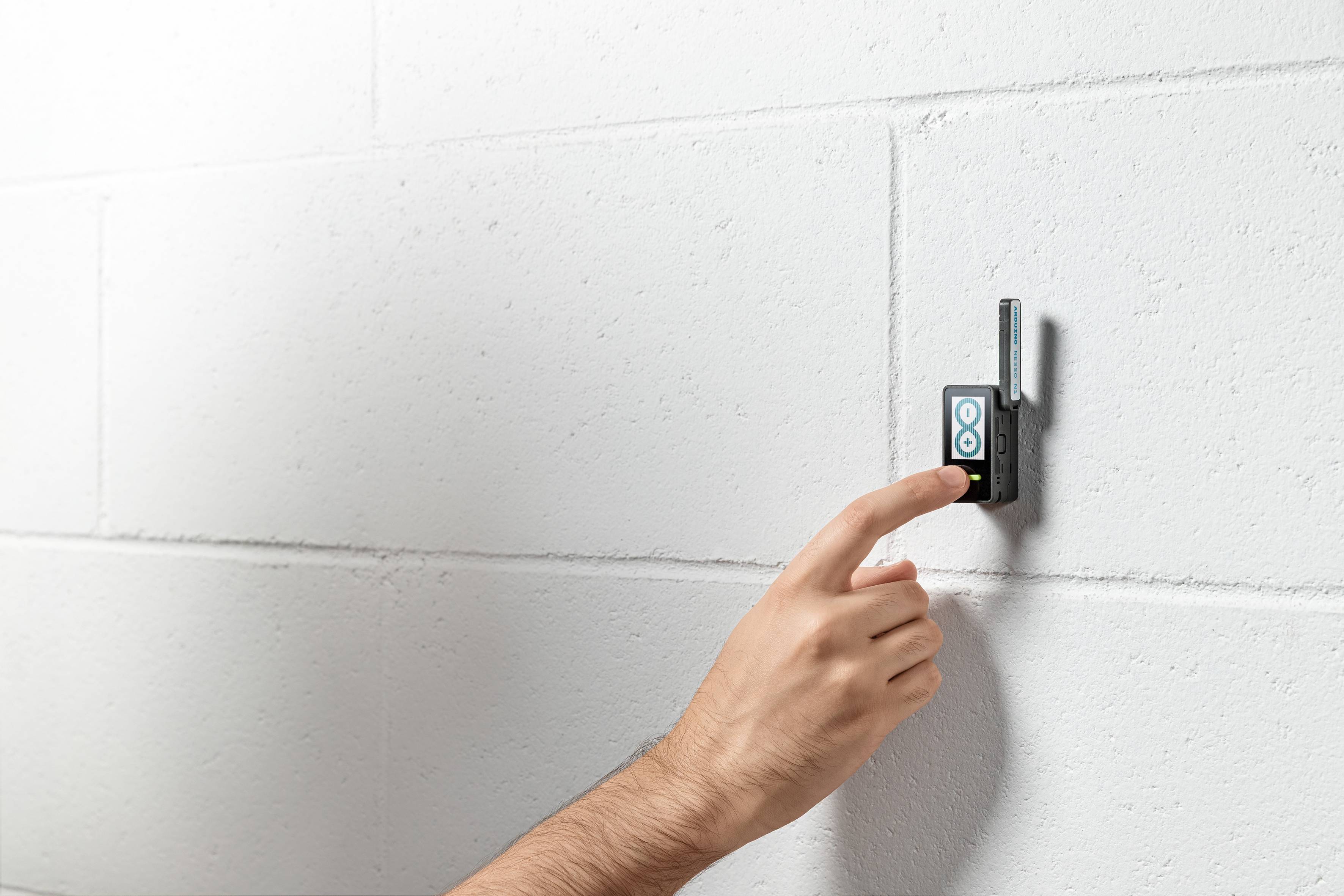 Eine Hand drückt einen Knopf auf einem kleinen schwarzen Gerät mit einer Antenne, das an einer weißen Ziegelwand montiert ist.