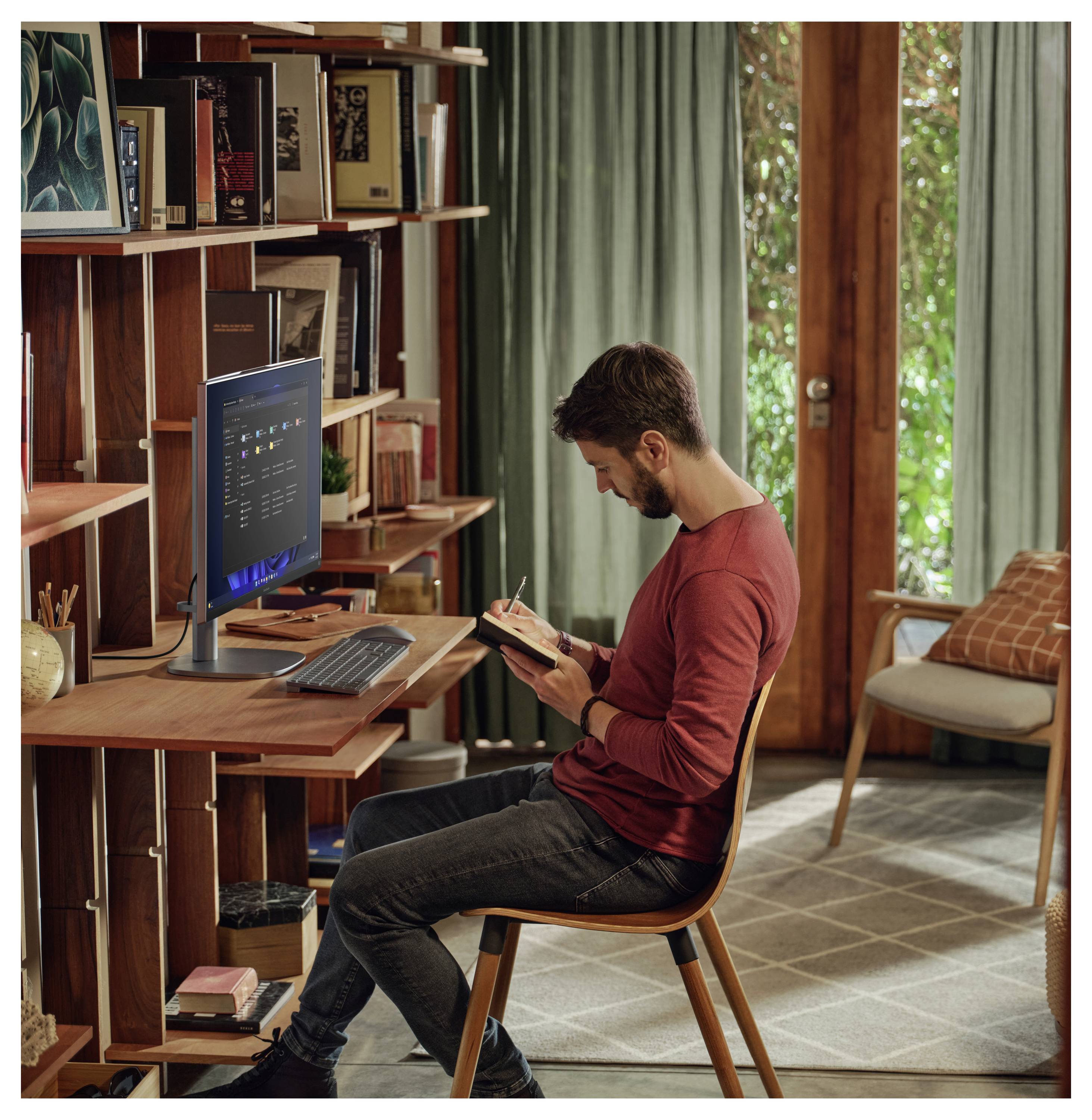 Ein Mann sitzt an einem Holzschreibtisch in einem Home Office und schreibt in ein Notizbuch. Ein Computerbildschirm zeigt geöffnete Anwendungen. Bücher und Dekoration stehen auf Regalen.