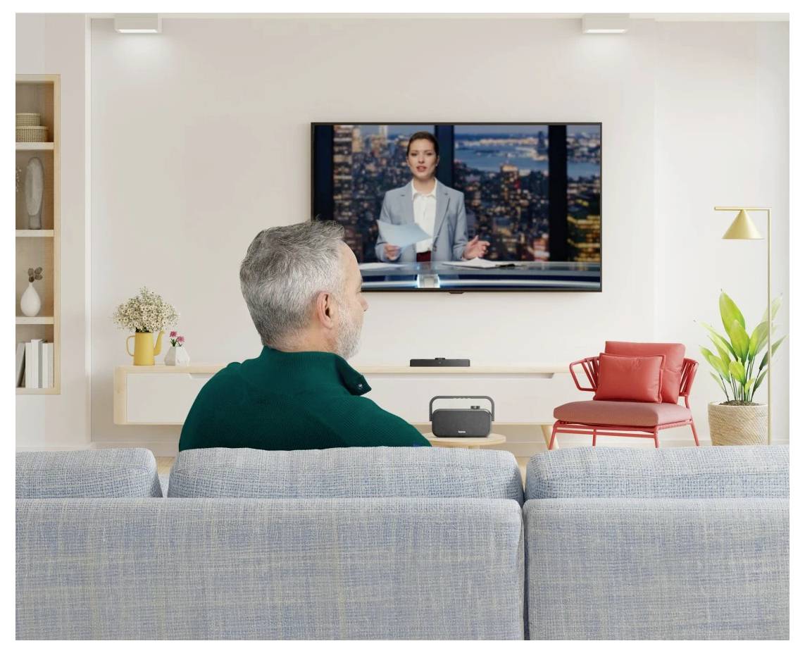 Ein älterer Mann verfolgt ein Fußballspiel im Fernsehen in einem modernen Wohnzimmer; ein Glas und ein Lautsprecher stehen auf der Anrichte vor ihm.