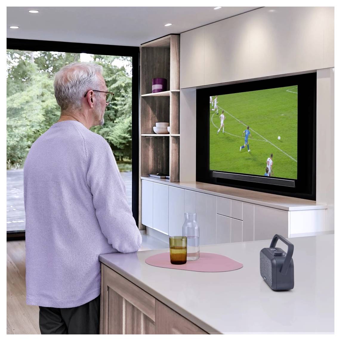 Ein älterer Mann schaut ein Fußballspiel auf einem großen Fernseher in einem modernen Wohnzimmer, mit einer malerischen Aussicht durch eine Glastür im Hintergrund.
