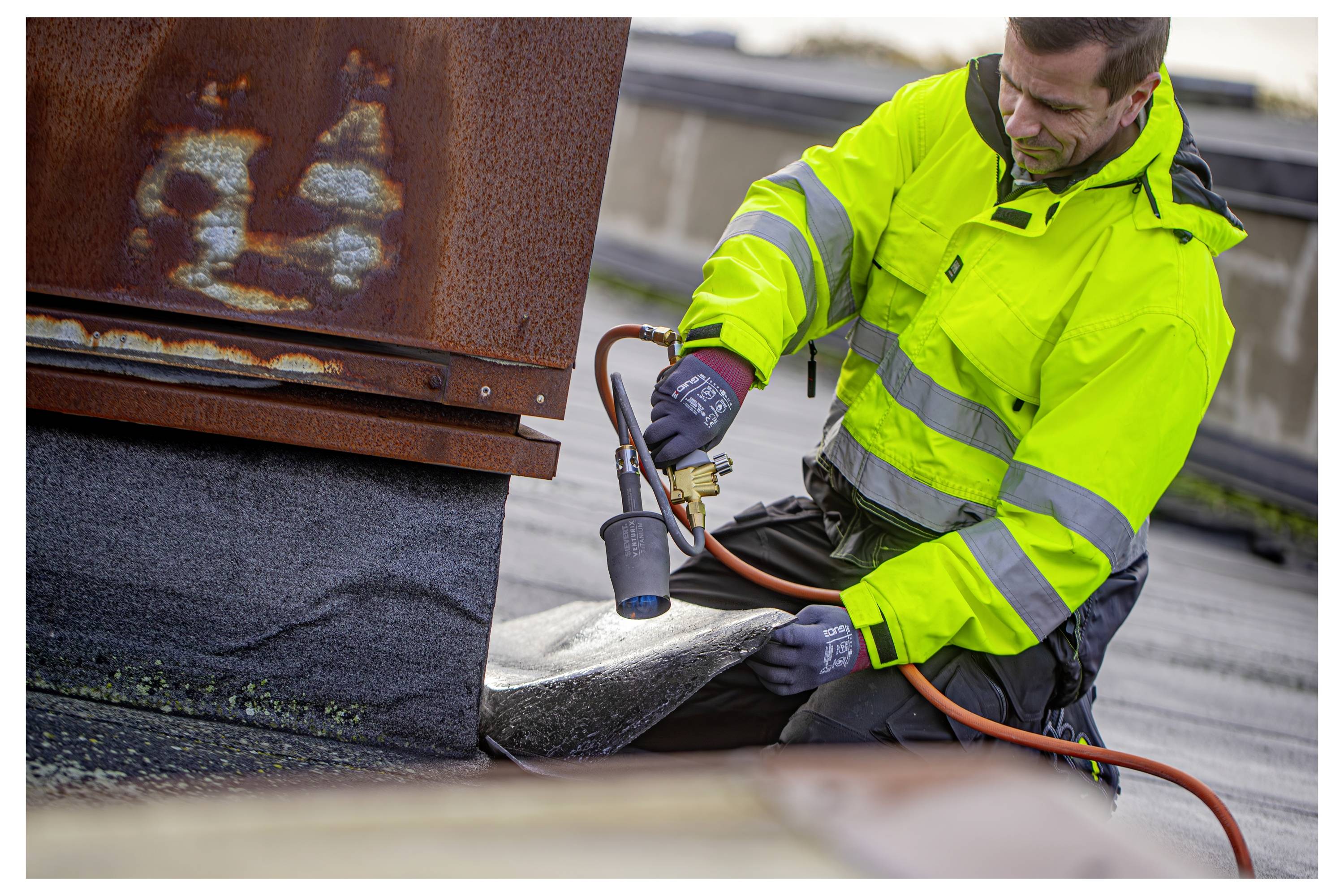 Ein Arbeiter in einer leuchtend gelben Jacke verwendet einen Brenner, um Dachmaterialien an einem Gebäude zu versiegeln und konzentriert sich dabei voll und ganz auf seine Aufgabe.