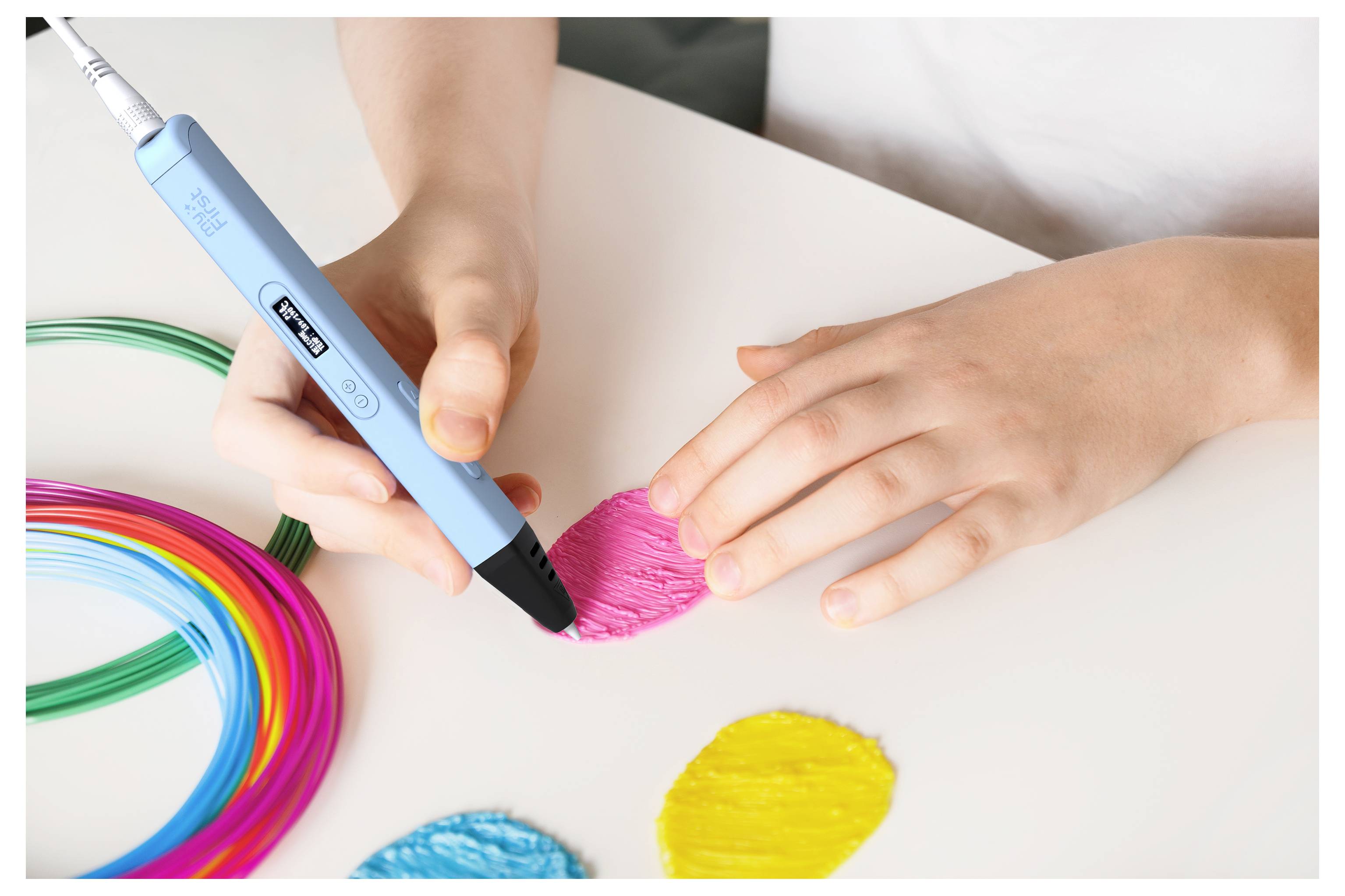 Eine Person verwendet einen blauen 3D-Stift, um eine rosa Form auf einem Tisch zu gestalten, wobei farbenfrohe Filamentspulen in der Nähe liegen.