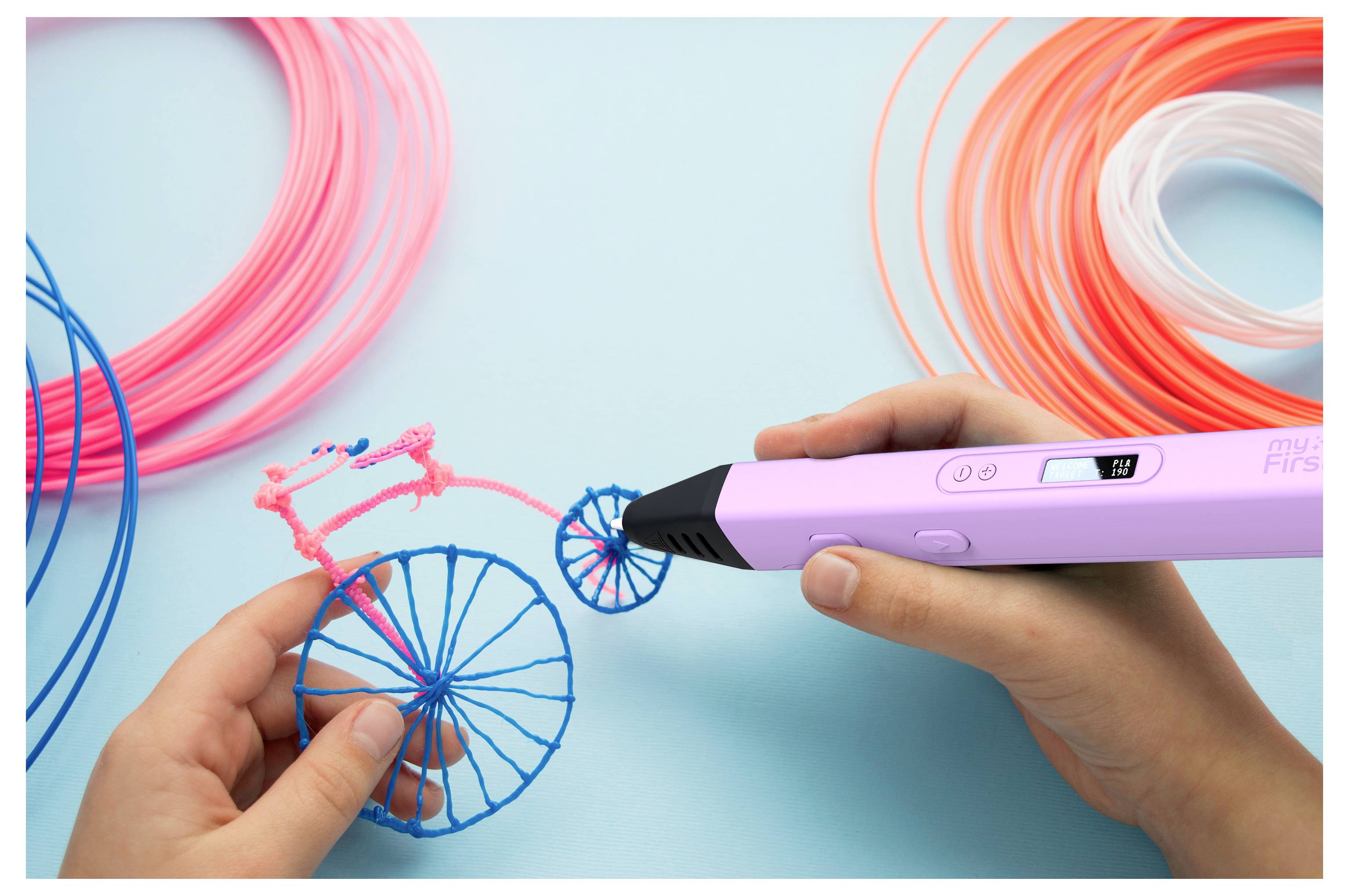 Hände, die einen 3D-Druckstift verwenden, erstellen ein kleines Fahrradmodell mit blauem und rosa Filament, umgeben von bunten Spulen auf einer hellblauen Oberfläche.