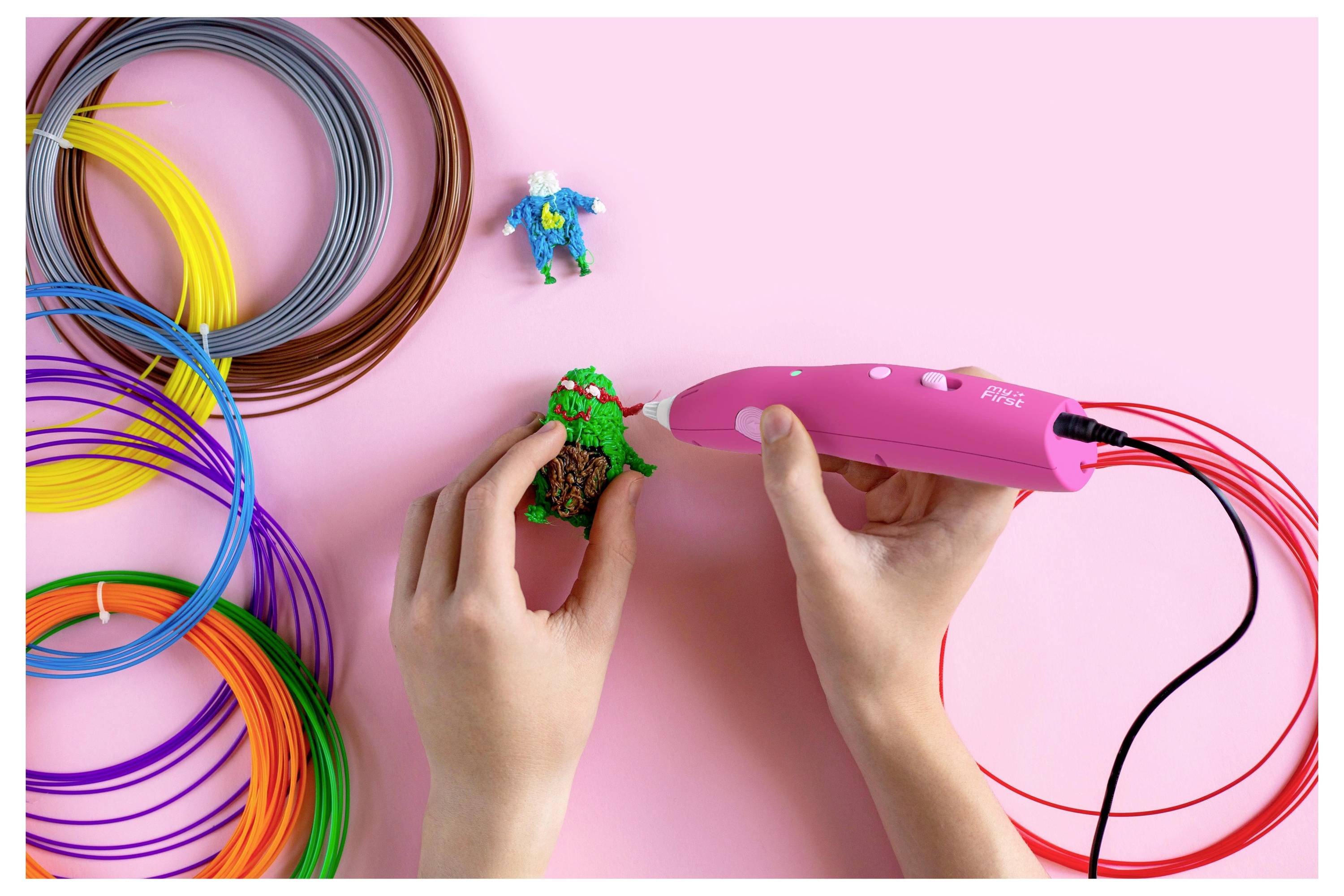 Zwei Hände verwenden einen rosa 3D-Stift, um ein grün-braunes Objekt zu erschaffen. Bunte Plastikfilamente und eine kleine blaue Figur befinden sich auf einem rosa Hintergrund.