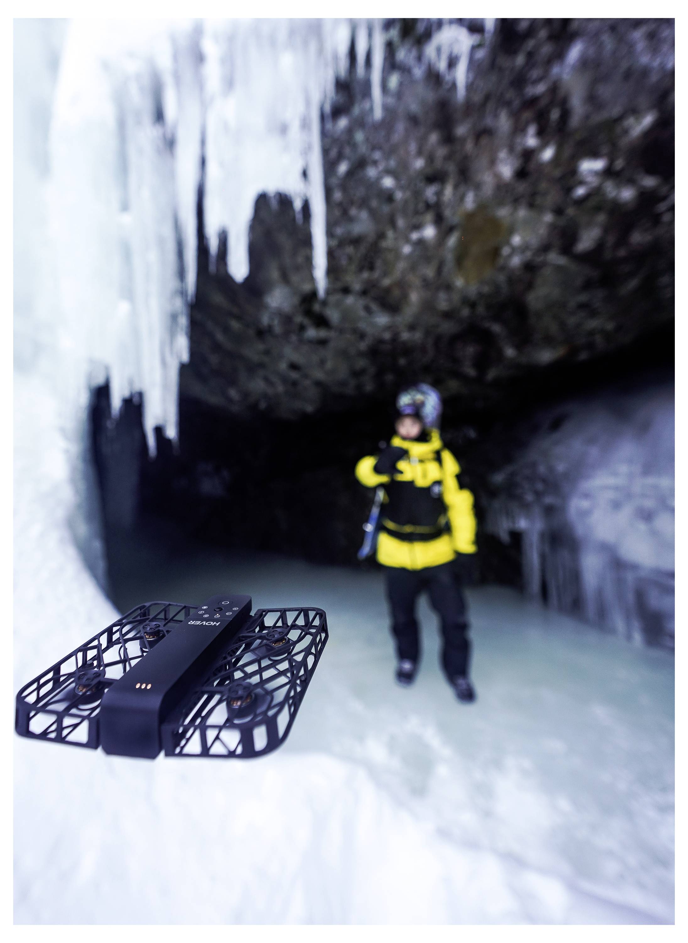 Eine Person in Winterausrüstung steht in einer eishöhle, mit Eiszapfen, die von der Decke hängen, während eine Drohne im Vordergrund schwebt.