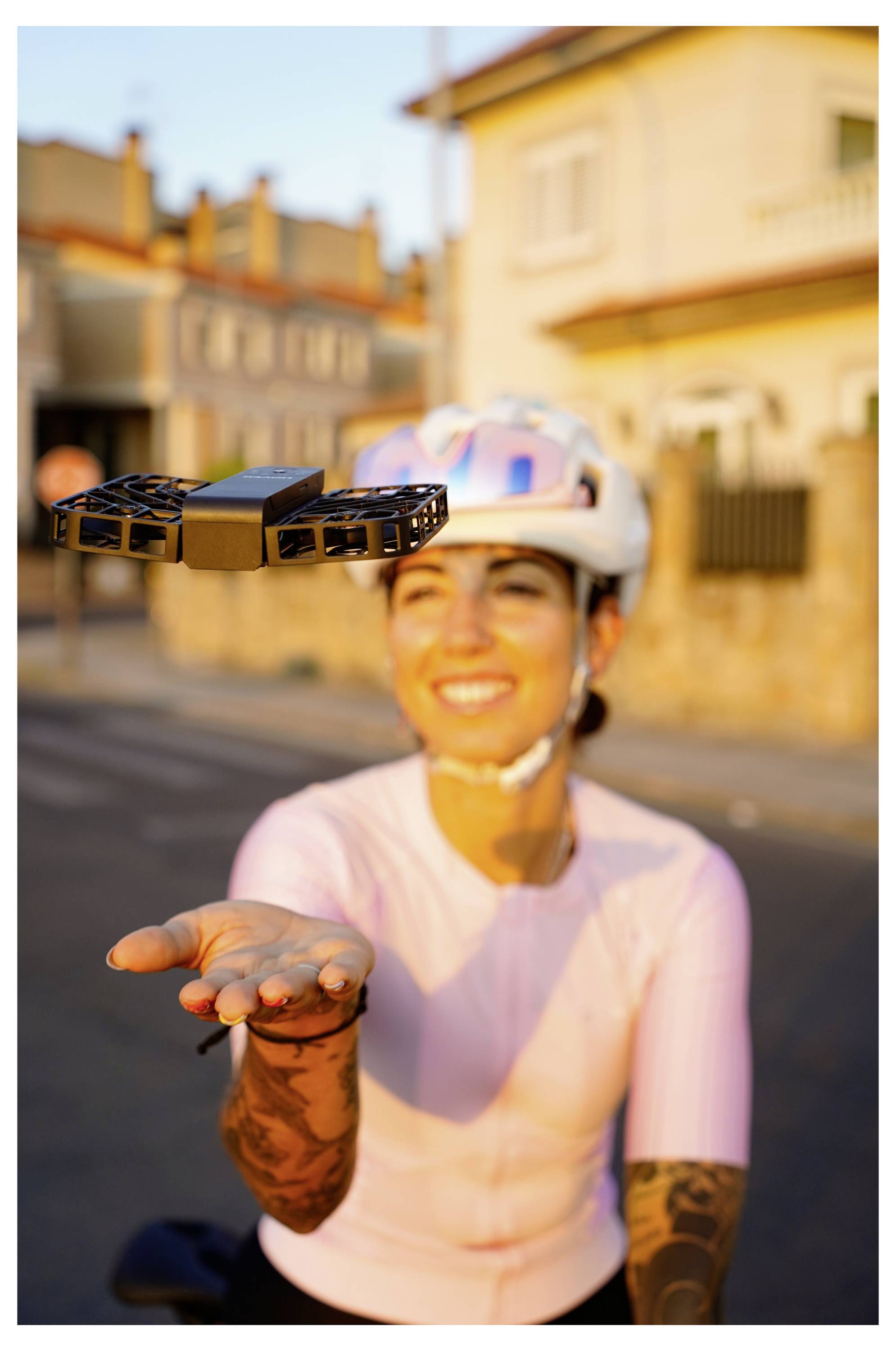 Eine Person mit Fahrradhelm und rosa Shirt lächelt, während sie an einem sonnigen Tag auf einer Straße einer schwebenden Drohne entgegengreift.