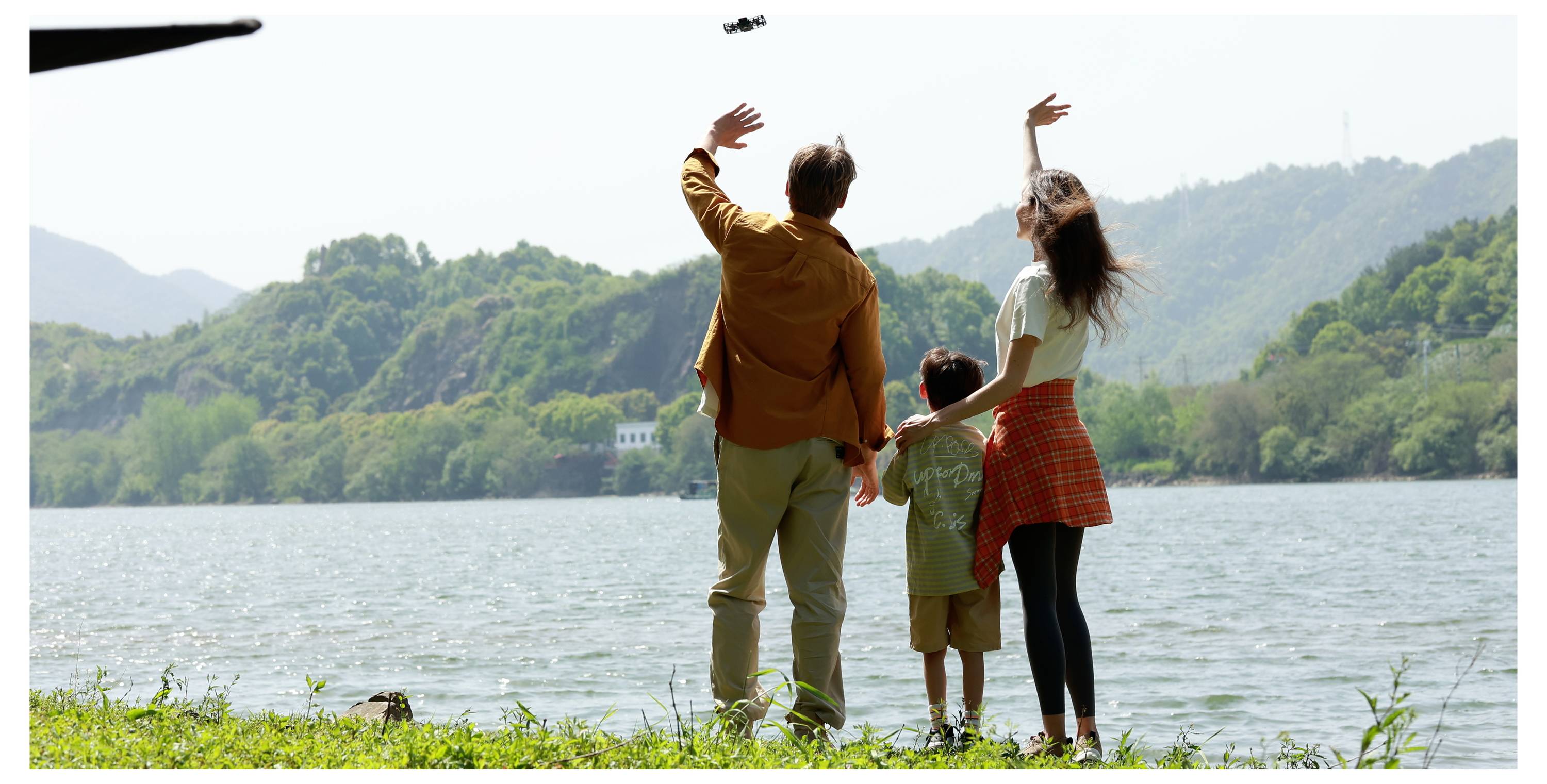 Eine Familie mit drei Personen steht an einem See und winkt einer Drohne, die über ihnen schwebt. Sie sind umgeben von üppiger Vegetation und fernen Hügeln unter einem klaren Himmel.