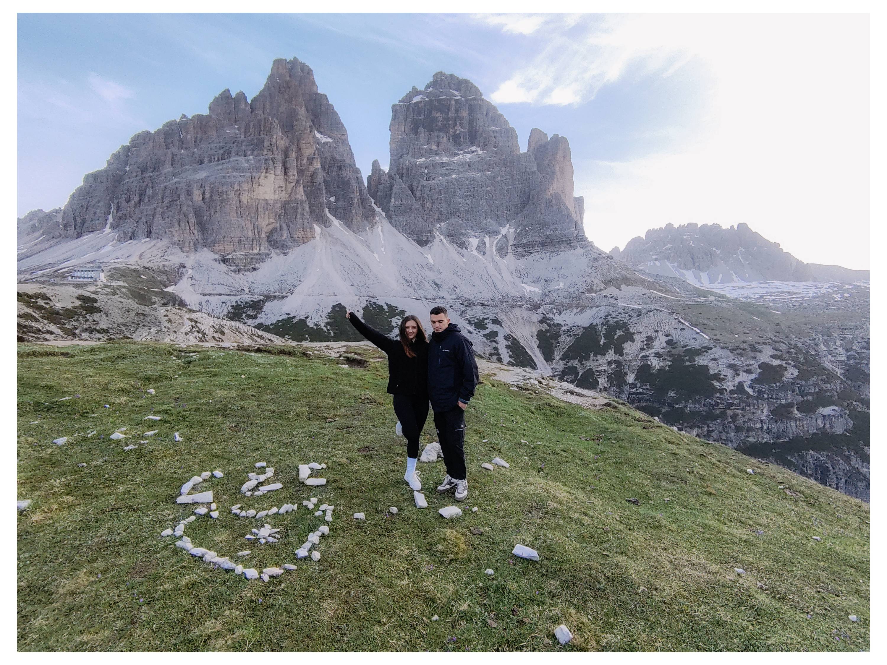 Ein Paar steht auf einem grasigen Hügel mit einer Herzform aus Steinen zu ihren Füßen. Majestätische Bergspitzen erheben sich im Hintergrund.