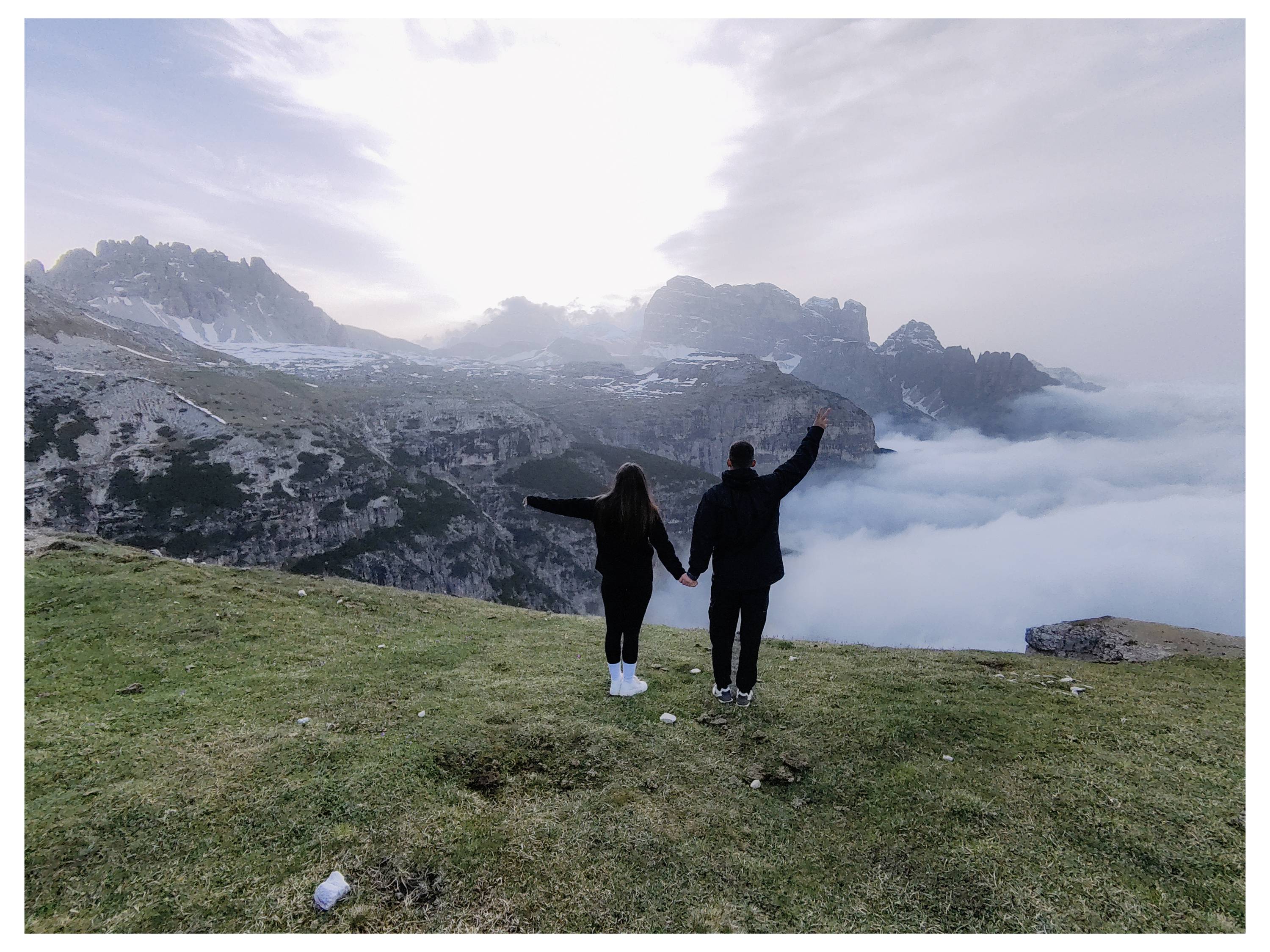 Ein Paar steht auf einem grasbewachsenen Hügel, hält Händchen und blickt auf eine malerische Aussicht mit Bergen und einem wolkengefüllten Tal unter einem strahlend blauen Himmel.