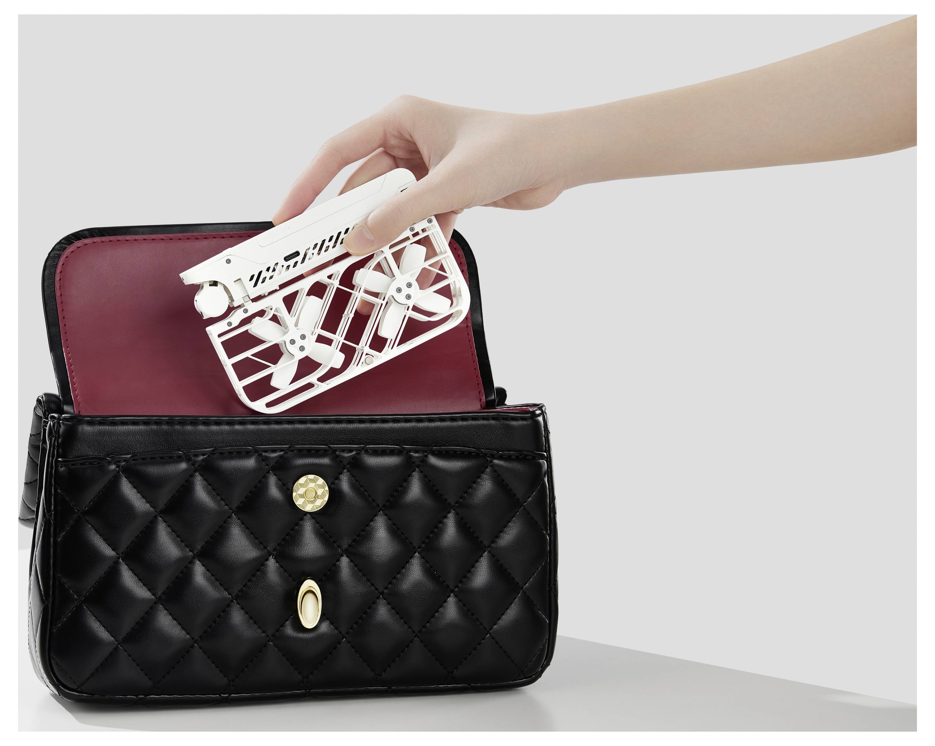 Eine Hand legt einen offenen weißen Fächer in eine schwarze gesteppte Handtasche mit goldenem Verschluss, vor einem einfarbigen Hintergrund.