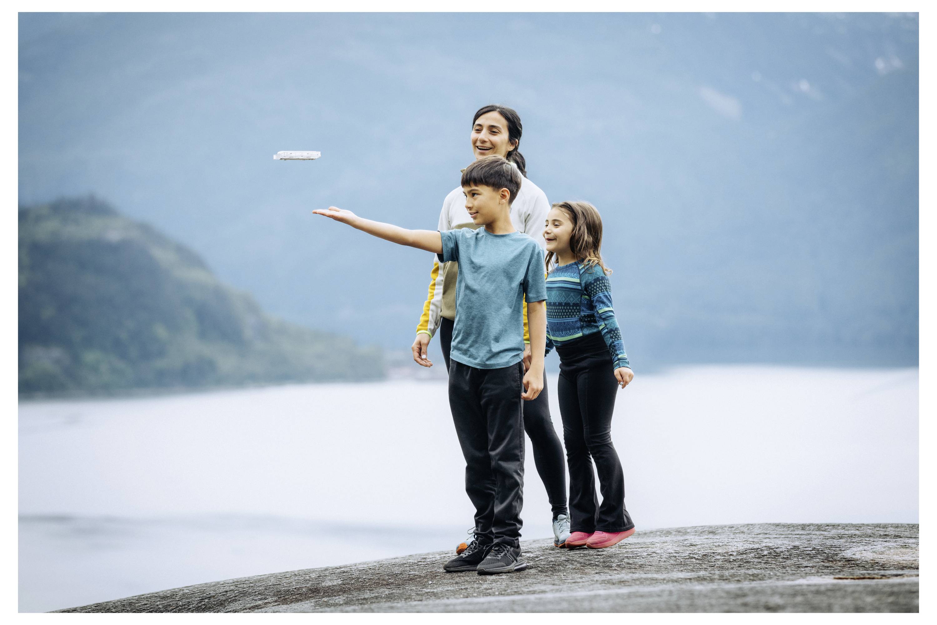Drei Kinder stehen auf einem Felsen an einem See mit nebligen Bergen im Hintergrund; ein Kind hat eine Drohne, die über seiner Handfläche schwebt.