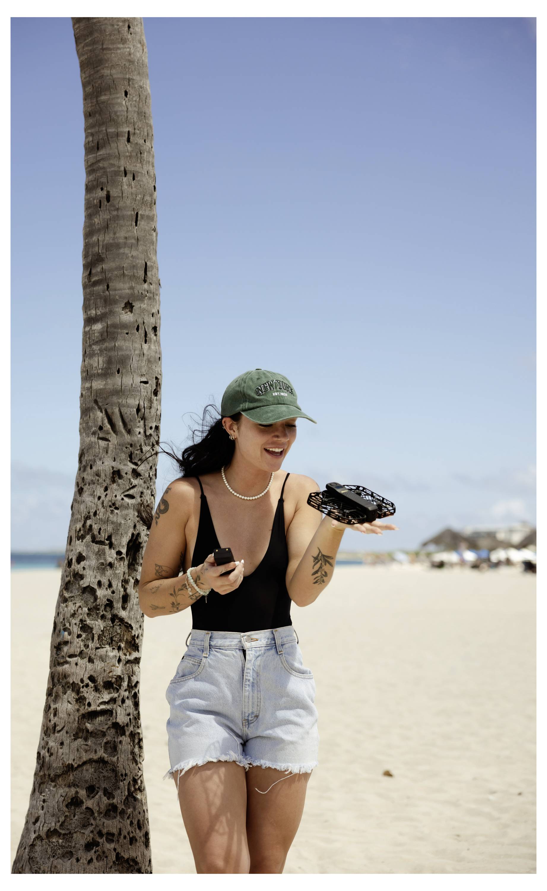 Eine Person mit grüner Kappe und legerer Kleidung lächelt neben einer Palme an einem sandigen Strand und hält eine Kamera. Im Hintergrund sind Strandhütten zu sehen.