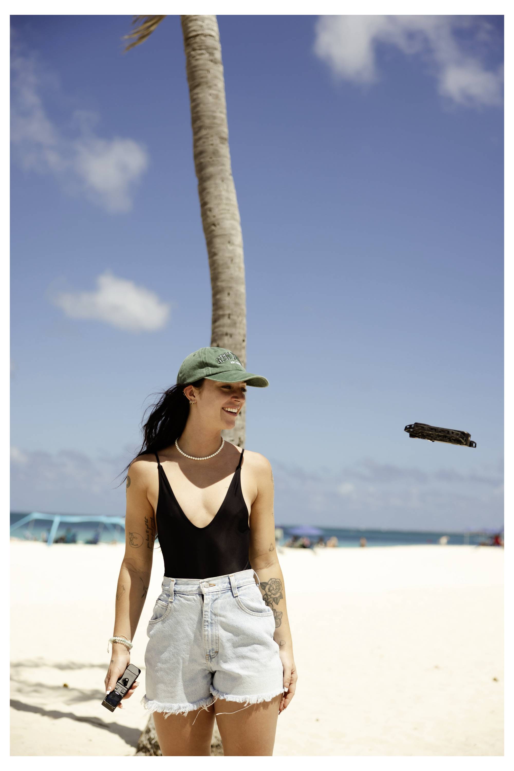Eine Person an einem sandigen Strand, die eine grüne Kappe, einen schwarzen Badeanzug und Jeansshorts trägt, mit Palmenbaum und blauem Himmel im Hintergrund.