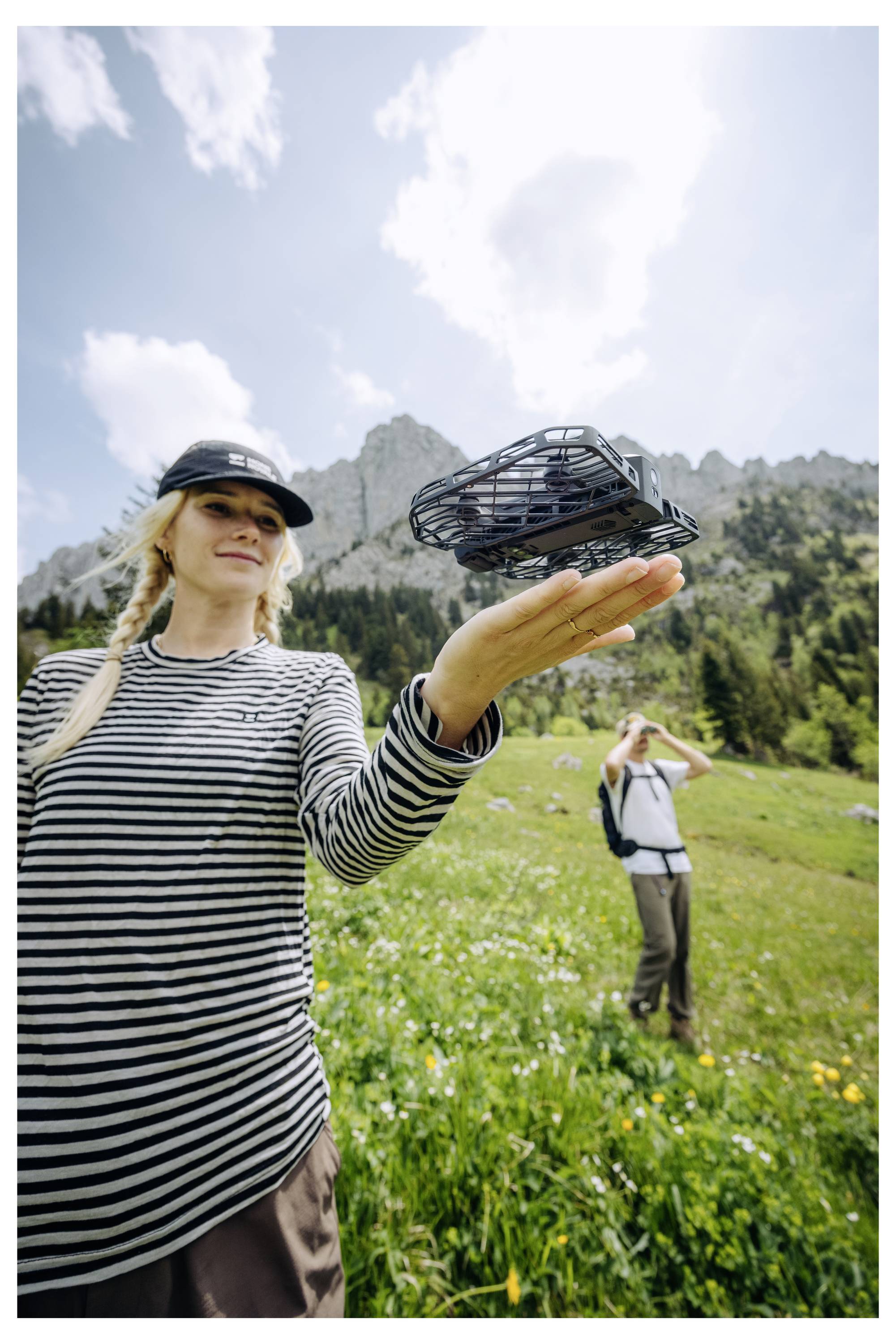 Frau, die eine kleine Drohne in einer grasigen Berglandschaft hält, während im Hintergrund eine Person ein Foto macht.