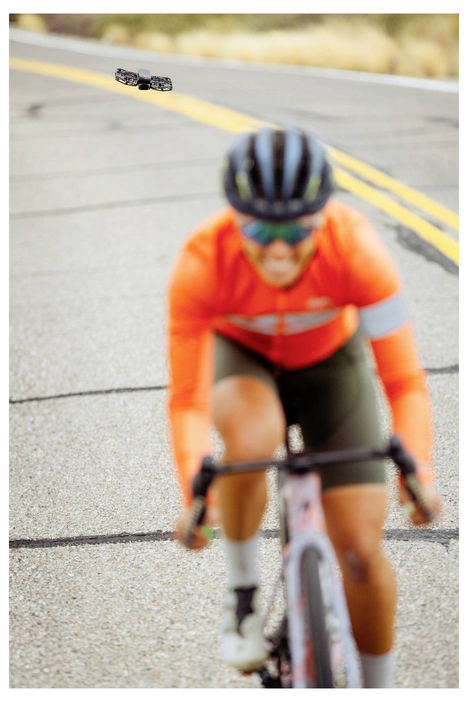Radfahrer in orangefarbener Jacke und grüner Hose, die auf einer Straße fährt, unscharfer Fokus, mit sichtbarem Pflaster und gelben Linien.