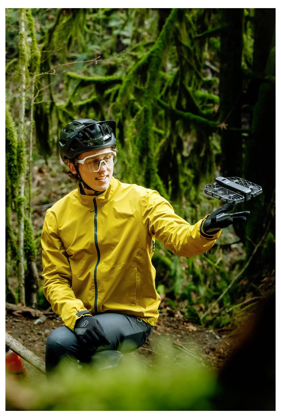 Eine Person in Outdoorkleidung, mit Helm und Handschuhen, hält ein kleines Gerät in einem bewaldeten Gebiet, umgeben von Grün.