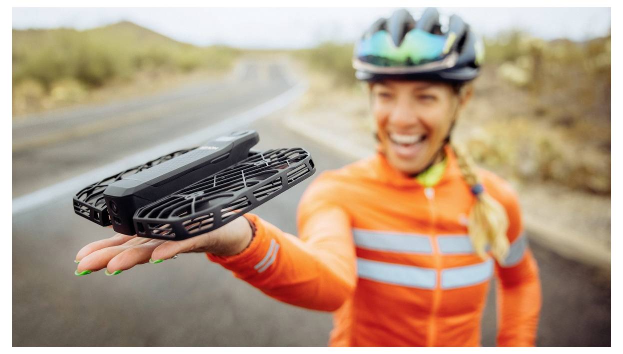 Ein Radfahrer in einer orangefarbenen Jacke hält eine kleine Drohne auf einer offenen Straße, lächelt und trägt einen Helm, mit einem verschwommenen Wüstenhintergrund.