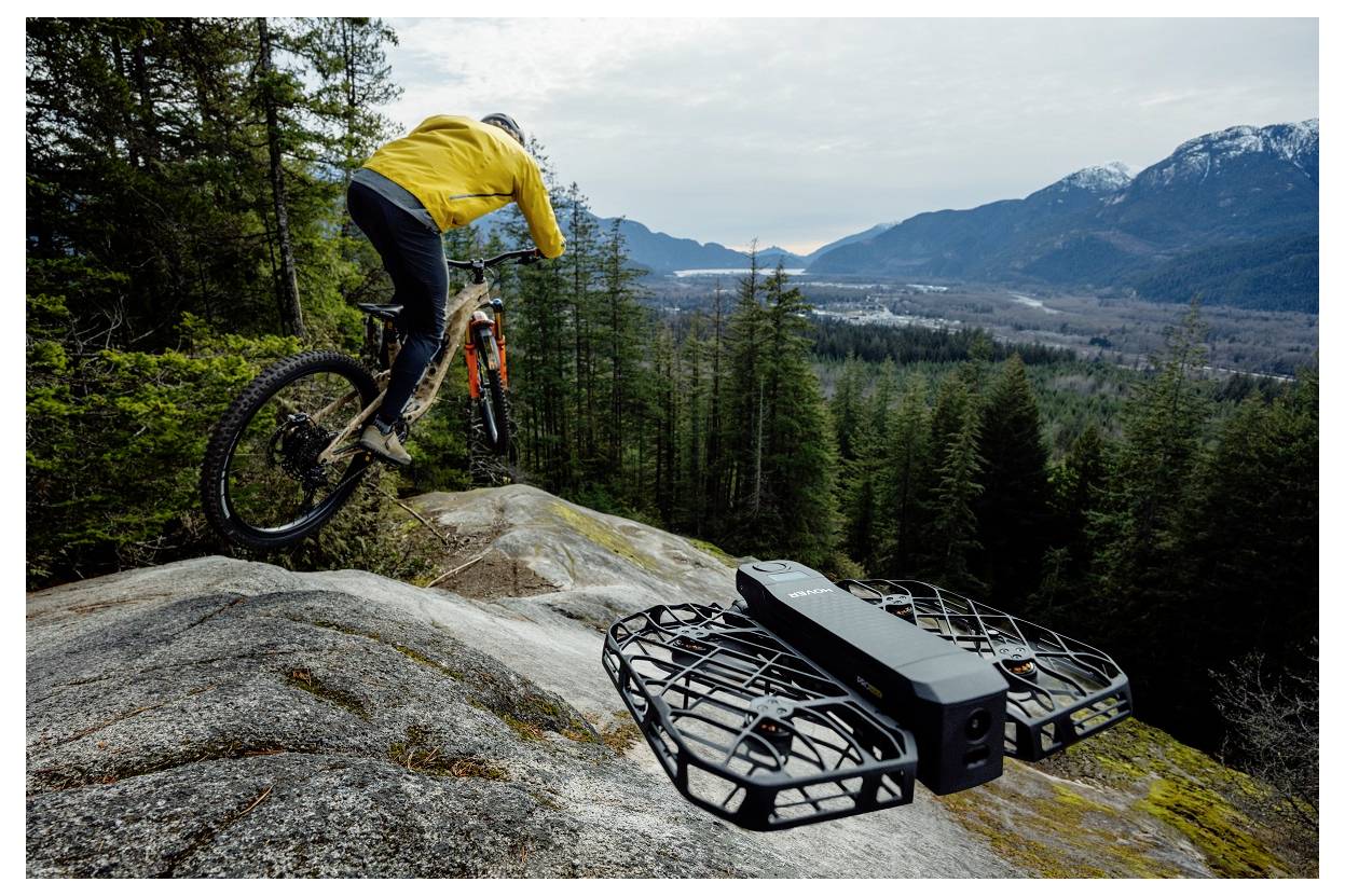 Eine Person in einer gelben Jacke fährt auf einem Mountainbike einen felsigen Pfad hinunter, während eine Drohne in der Nähe schwebt, vor einer Kulisse von bewaldeten Hügeln.