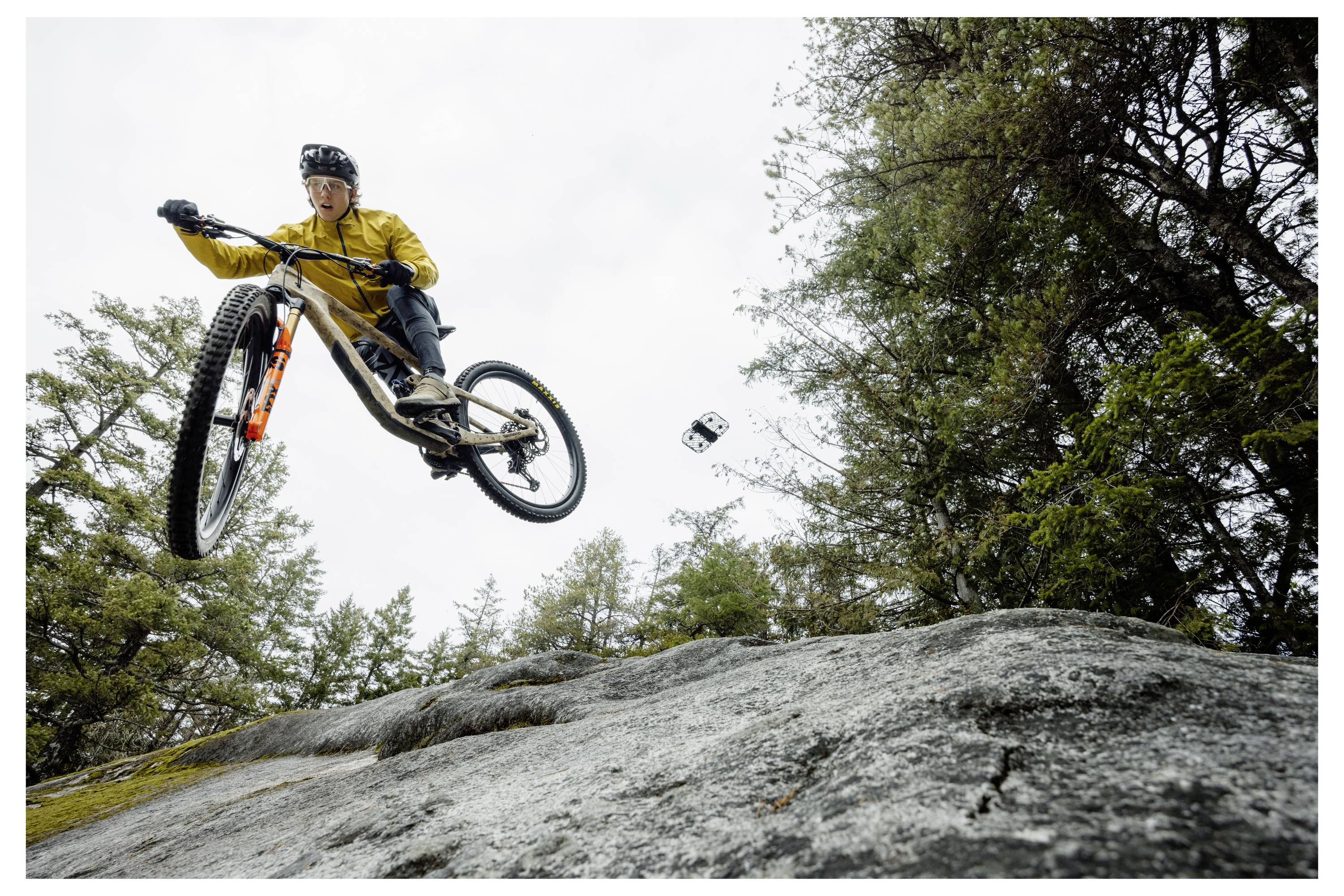 Eine Person mit einer gelben Jacke und einem Helm führt einen Sprung auf einem Mountainbike von einer felsigen Oberfläche in einem bewaldeten Gebiet durch.