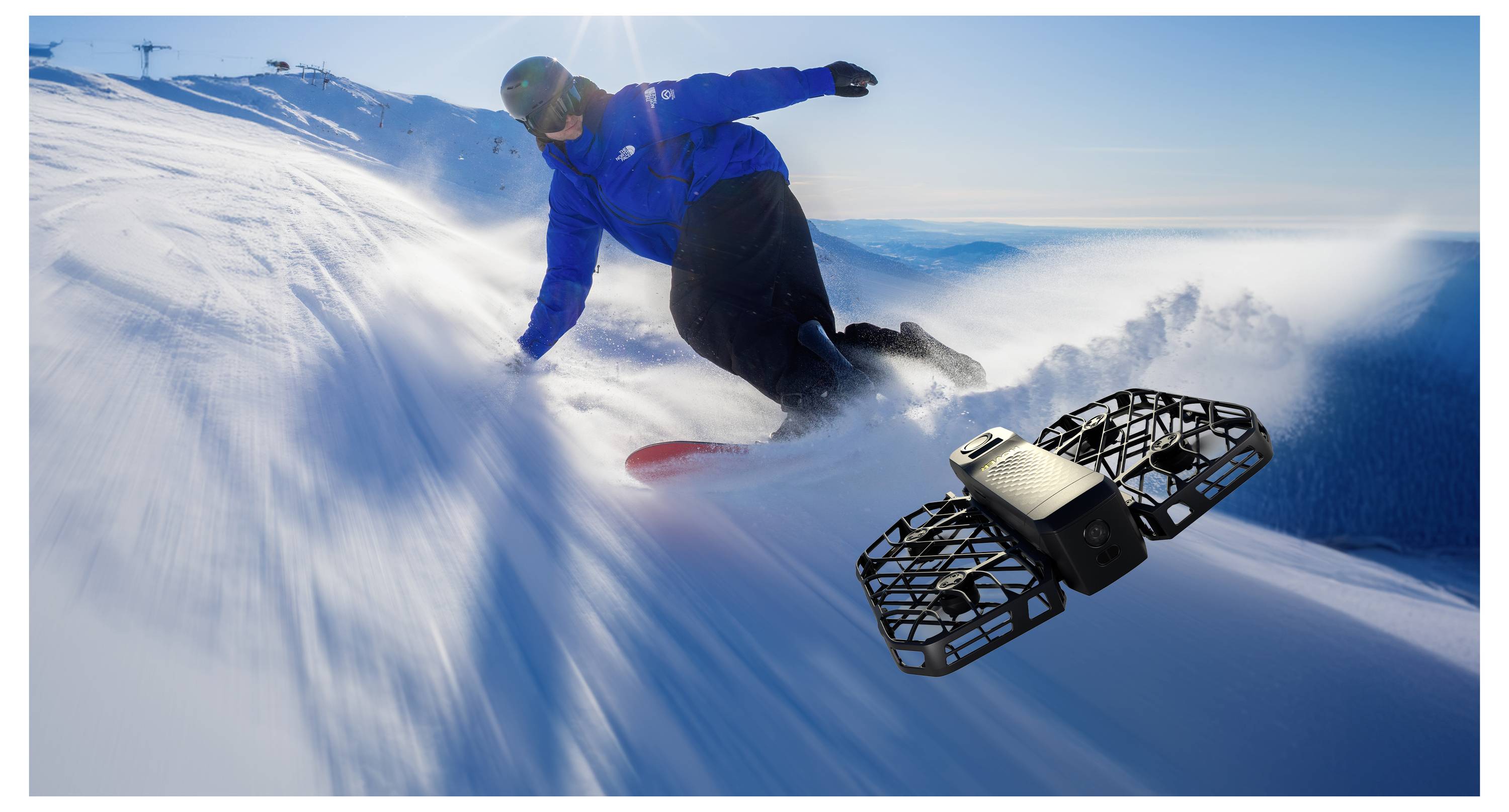 Eine Person in einer blauen Jacke fährt auf einem schneebedeckten Berghang Snowboard bergab, gefolgt von einer kleinen Drohne, die die Aktion aufnimmt.