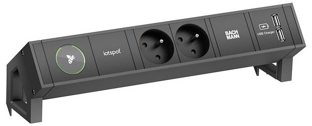 Eine schwarze Steckdosenleiste mit zwei Steckdosen, einem USB-Ladegerät, einem „BACHMANN"-Logo und einer „iotspot"-Kontrolltaste mit grünem Ring.