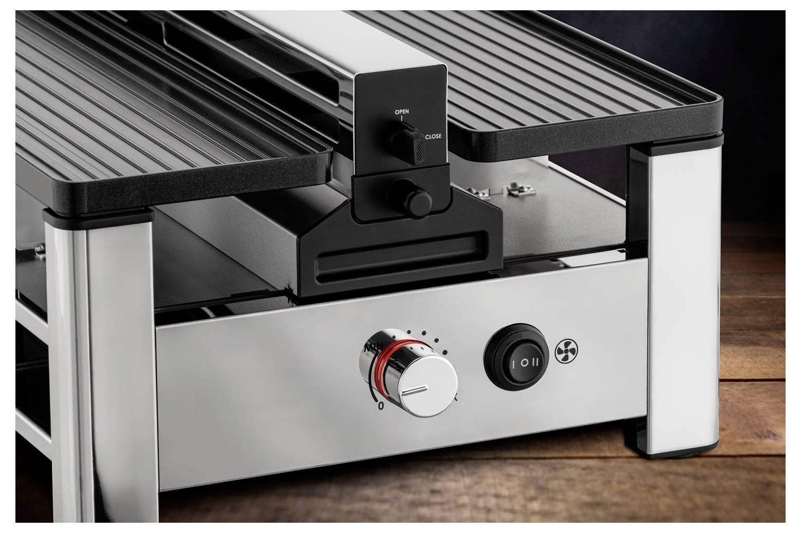 Ein silberner, elektrischer Grill mit Drehknopf und Netzschalter auf einem Holztisch. Das Design ist modern und funktional.