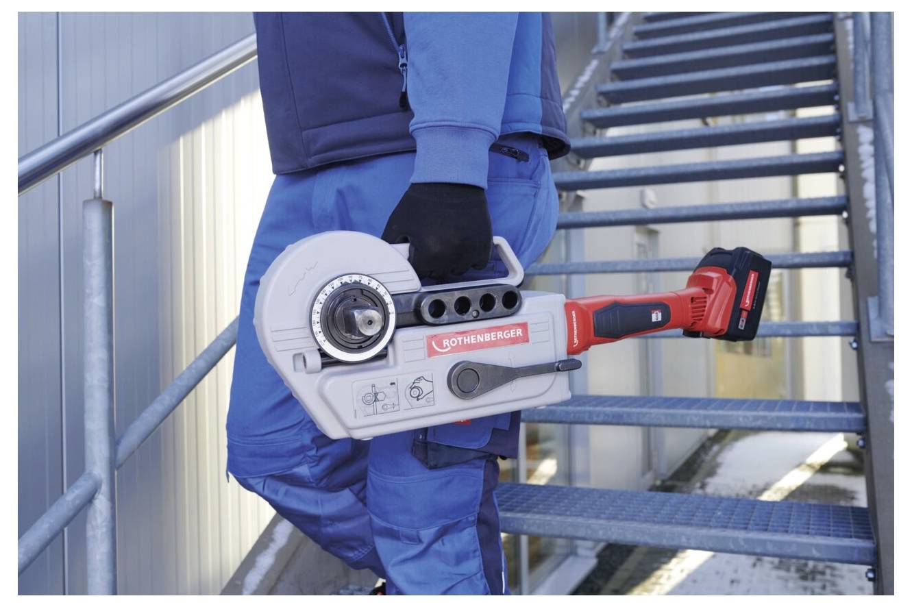 Person in blauer Arbeitskleidung trägt ein großes rotes und silbernes Werkzeug, möglicherweise eine Rohrzange, während sie industrielle Treppe hinaufgeht.