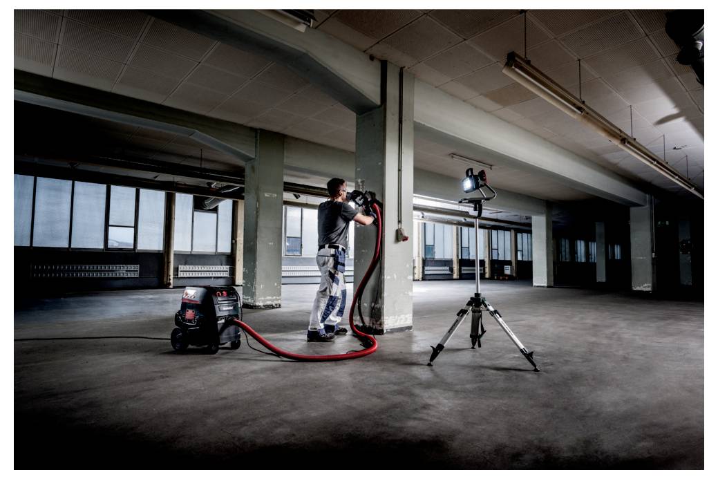 Eine Person in Arbeitskleidung bedient einen industriellen Staubsauger in einem großen, leeren Industrieraum, wobei eine auf einem Stativ montierte Lampe auf den Arbeitsbereich gerichtet ist.