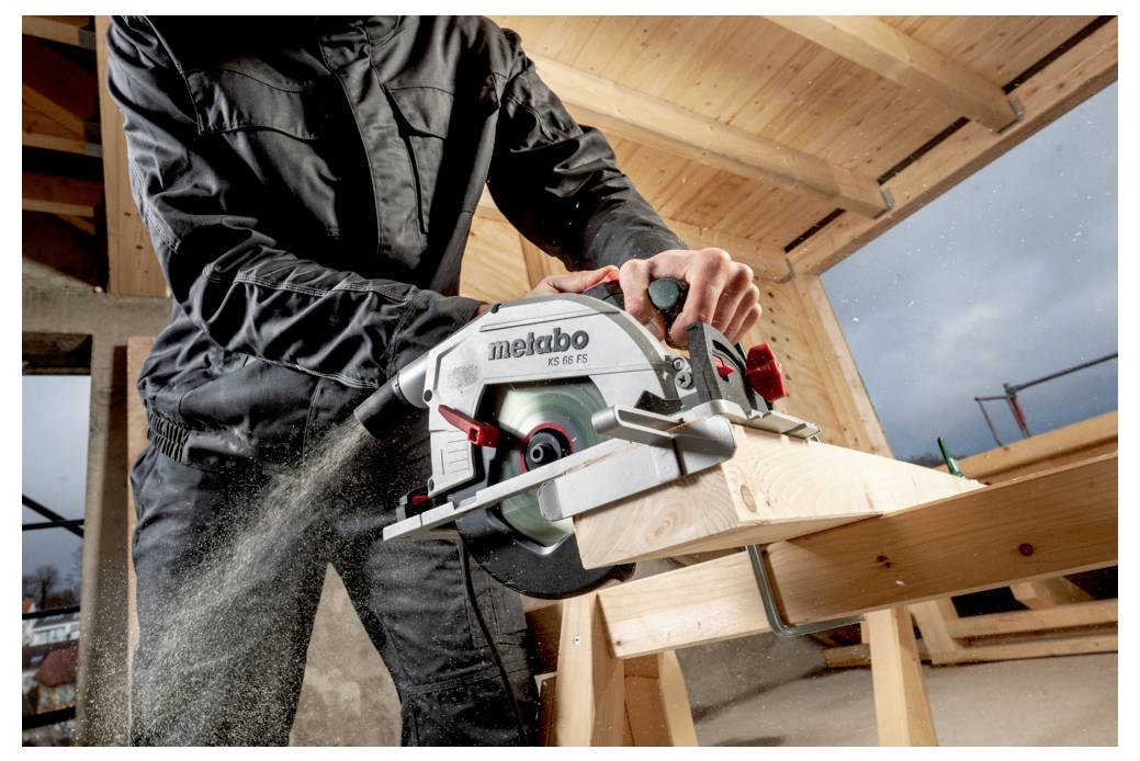 Person in einer Werkstatt, der eine Holzplanke mit einer Handkreissäge zersägt und dabei Sägespäne produziert, unter einem teilweise fertiggestellten Holzdach.