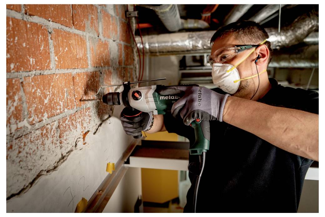 Eine Person mit Mundschutz und Handschuhen bedient eine Bohrmaschine an einer Ziegelwand, umgeben von Baumaterialien und Rohrleitungen.