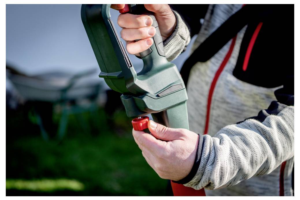 Eine Person dreht an einem roten Knopf an einem grünen Gartengerät, möglicherweise einem Laubgebläse, im Freien in einer Gartenumgebung.