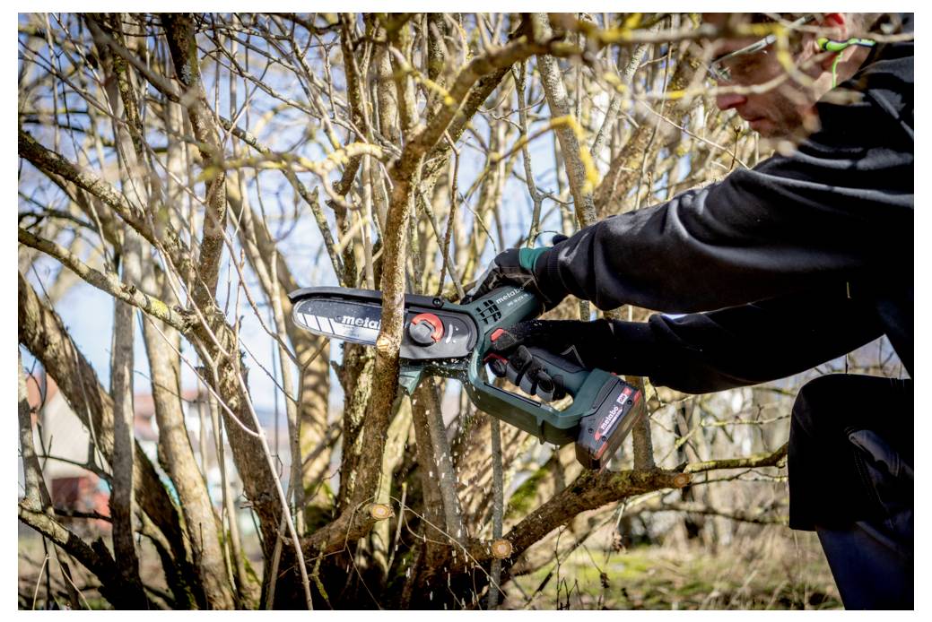 Eine Person verwendet eine kleine elektrische Kettensäge, um Äste an einem Baum oder Strauch in einem Garten zu beschneiden und trägt dabei Schutzausrüstung.