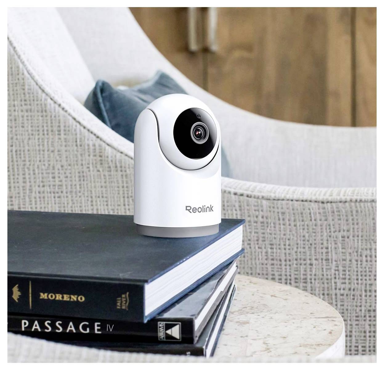 Eine Reolink-Sicherheitskamera steht auf einem Stapel von Büchern mit den Titeln "Passage IV" und "Moreno", der auf einem Beistelltisch neben einem gepolsterten Stuhl platziert ist.