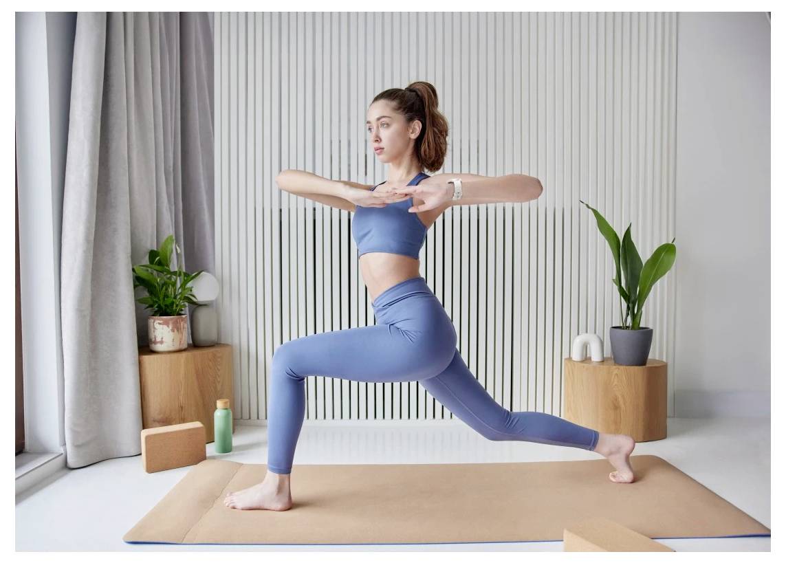 Eine Frau in einem blauen Trainingsanzug führt einen Ausfallschritt auf einer Yogamatte in einem hellen Raum mit Pflanzen und Trainingsutensilien durch.