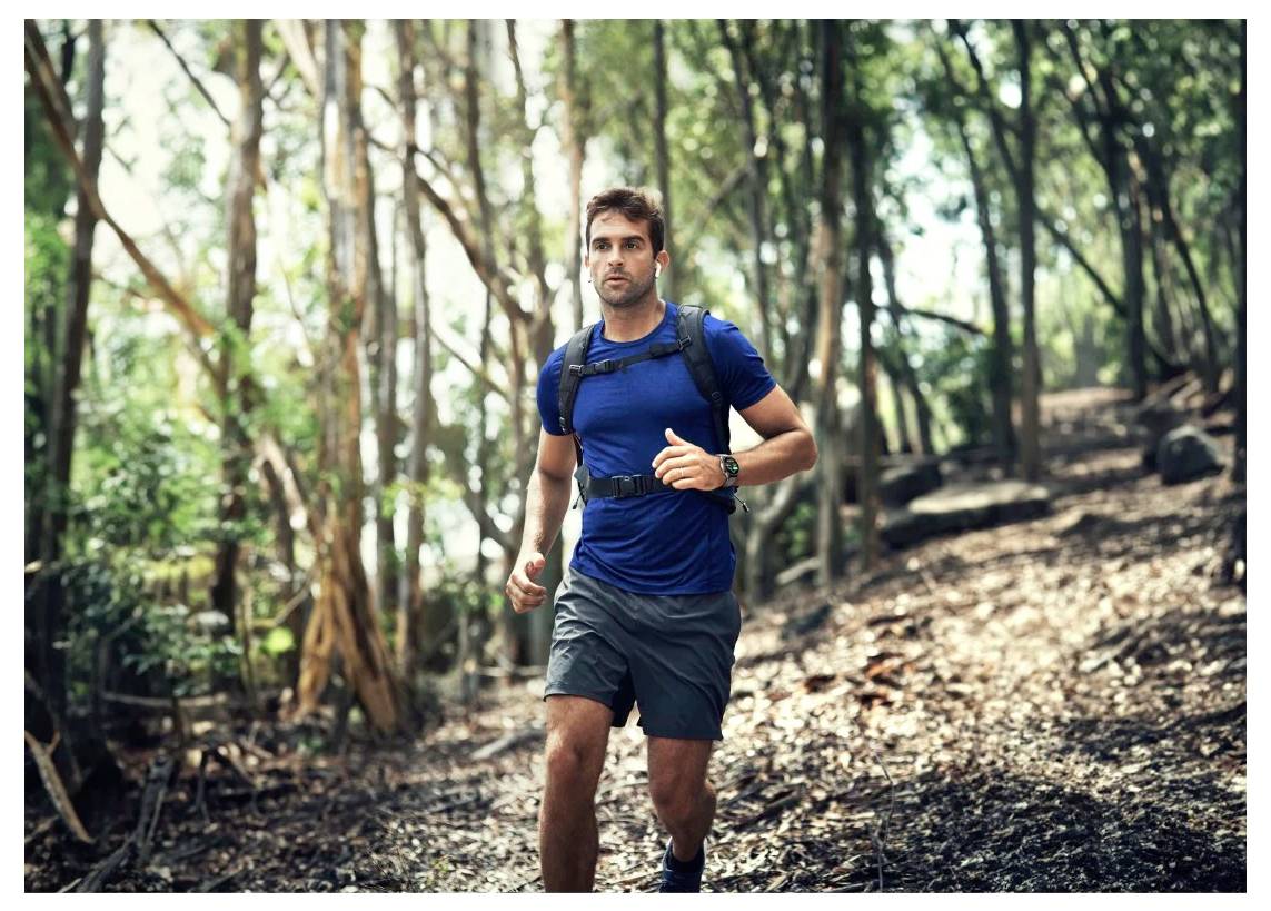 Eine Person, die einen Waldweg entlang joggt, trägt ein blaues Shirt und hat einen kleinen Rucksack dabei, umgeben von hohen Bäumen und Sonnenlicht.