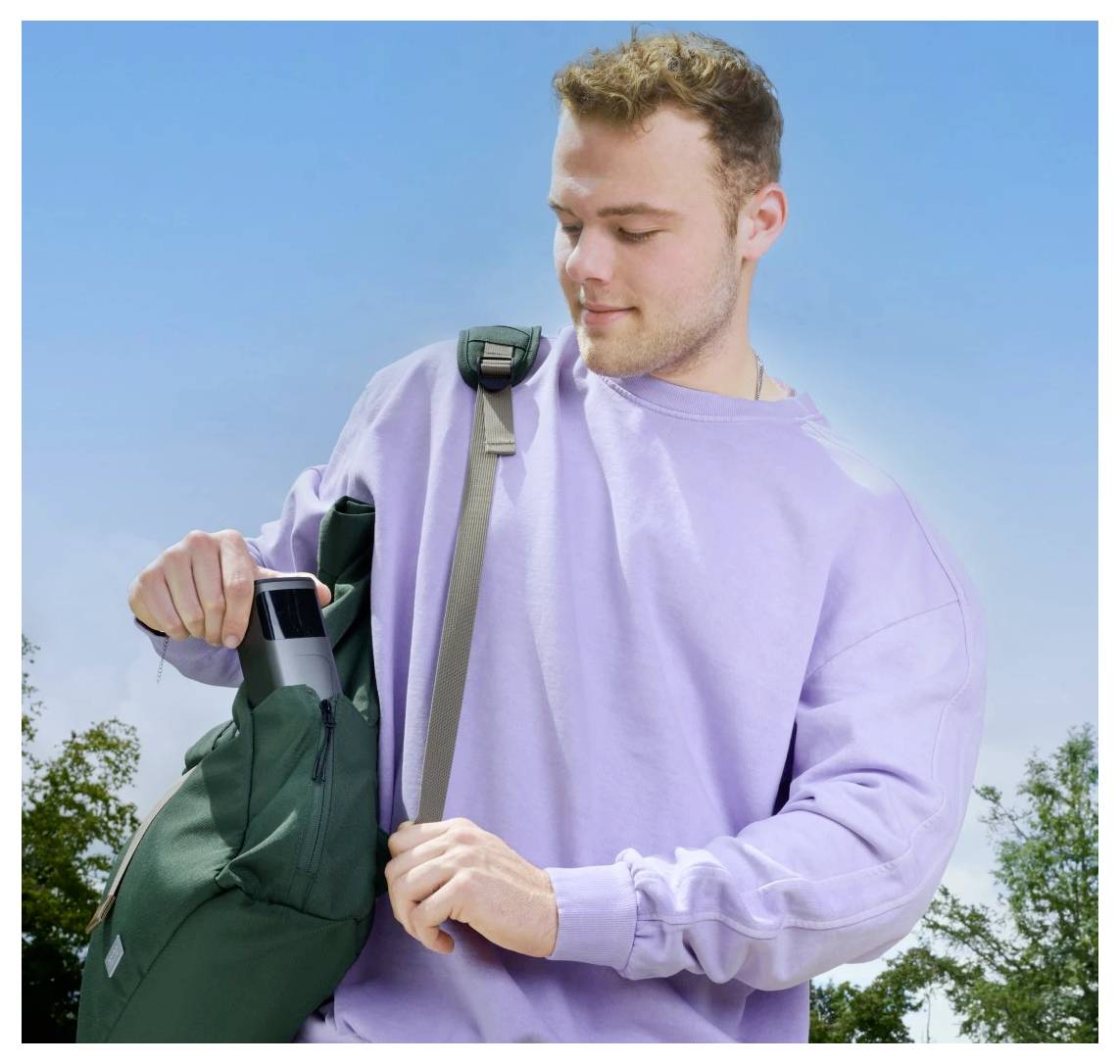 Eine Person in einem lila Sweatshirt legt eine schwarze Wasserflasche in einen grünen Rucksack vor einem klaren blauen Himmelshintergrund.