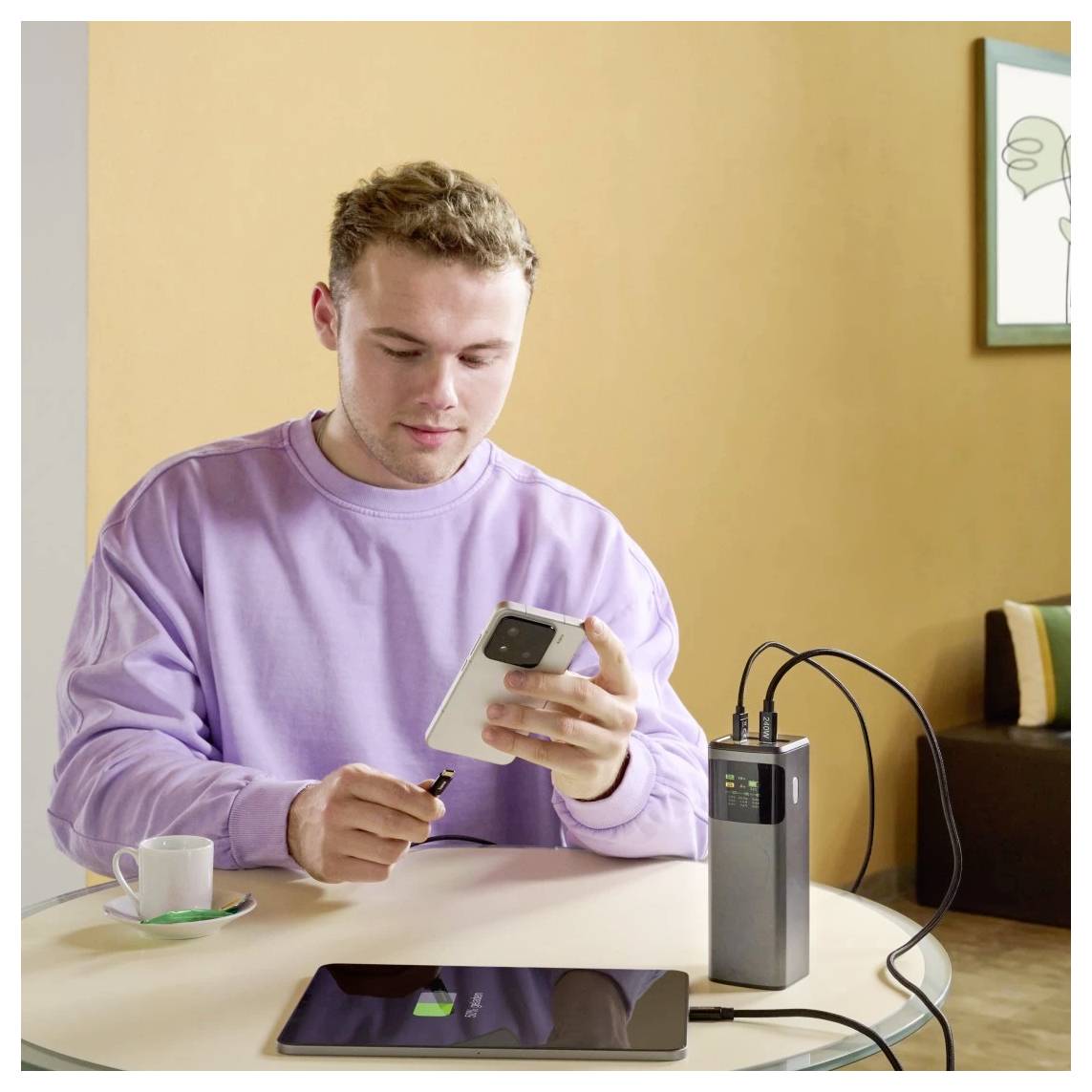 Eine Person schließt ein Smartphone an einer Ladestation an, sitzt an einem Tisch mit einer Kaffeetasse in der Nähe. Im Hintergrund ist ein Sofa zu sehen.