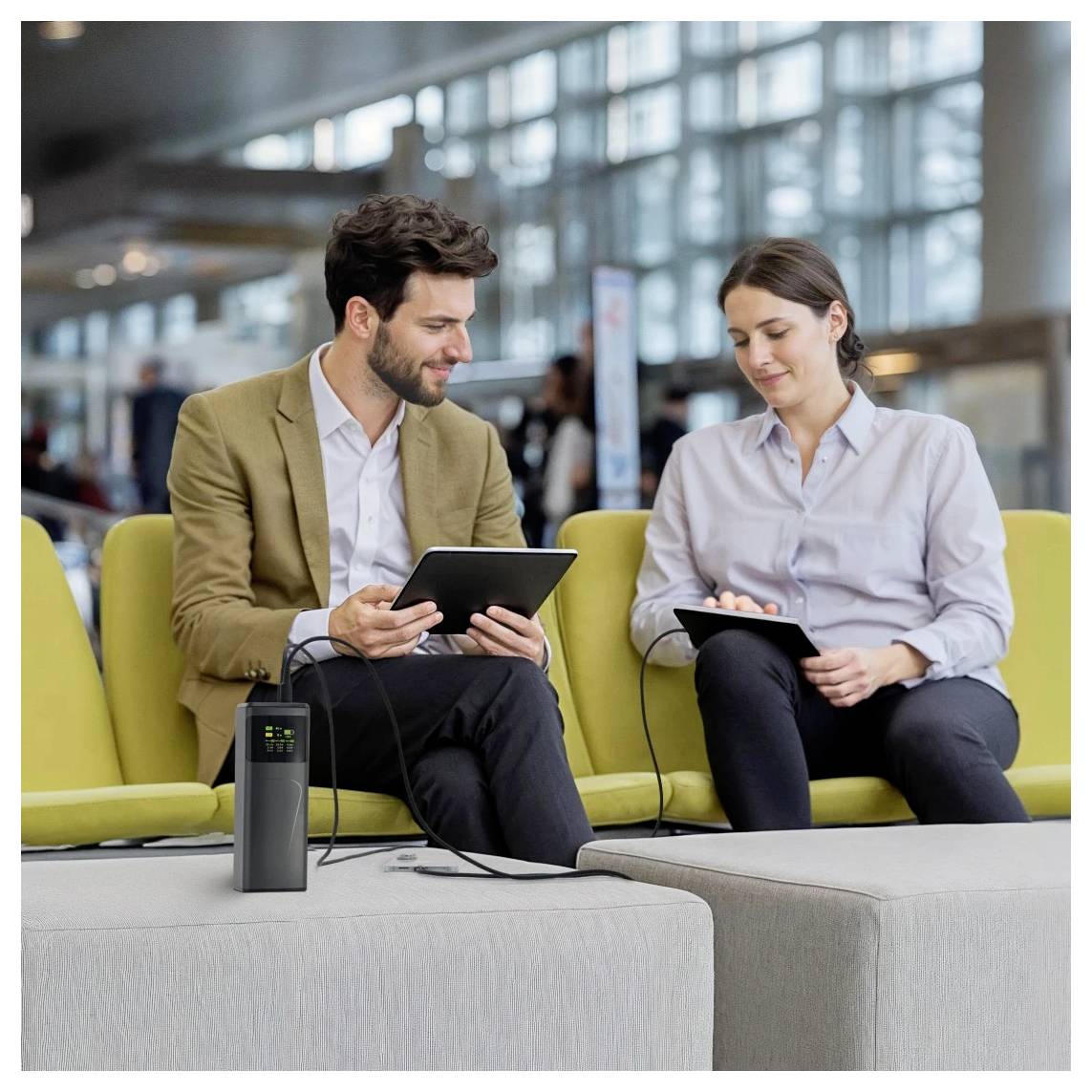 Ein Mann und eine Frau sitzen auf einer Bank in einer modernen Umgebung, beide nutzen Tablets, die mit einer mobilen Stromstation auf einem Tisch verbunden sind.
