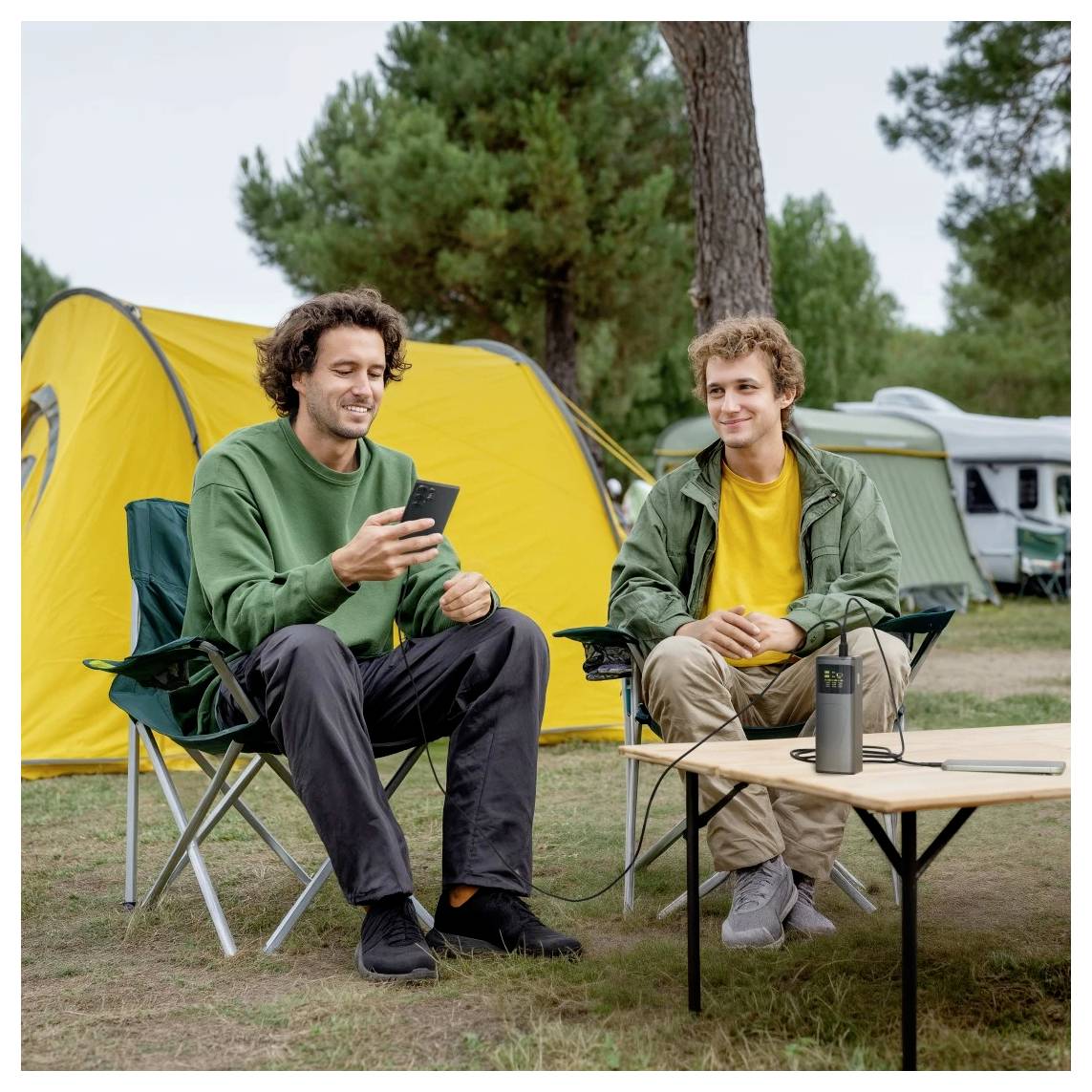 Zwei Personen sitzen in Campingstühlen neben einem gelben Zelt, wobei eine Person ein Smartphone benutzt. Eine Powerbank auf dem Tisch deutet auf eine technikfreundliche Einrichtung hin.