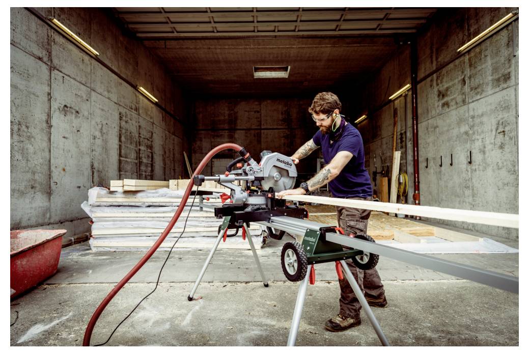 Eine Person in einer Werkstatt schneidet Holz mit einer Kreissäge. Der Raum ist industriell mit Betonwänden und gestapelten Materialien. Schutzausrüstung wird getragen.