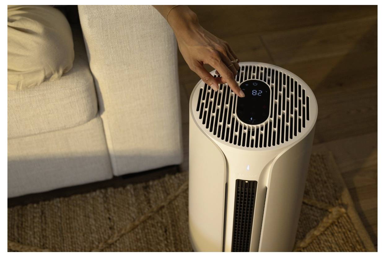 Eine Person stellt die Temperatur an einem kompakten Luftreiniger ein, der eine '82' auf einem digitalen Bildschirm anzeigt und auf einem strukturierten Teppich in der Nähe eines Sofas platziert ist.