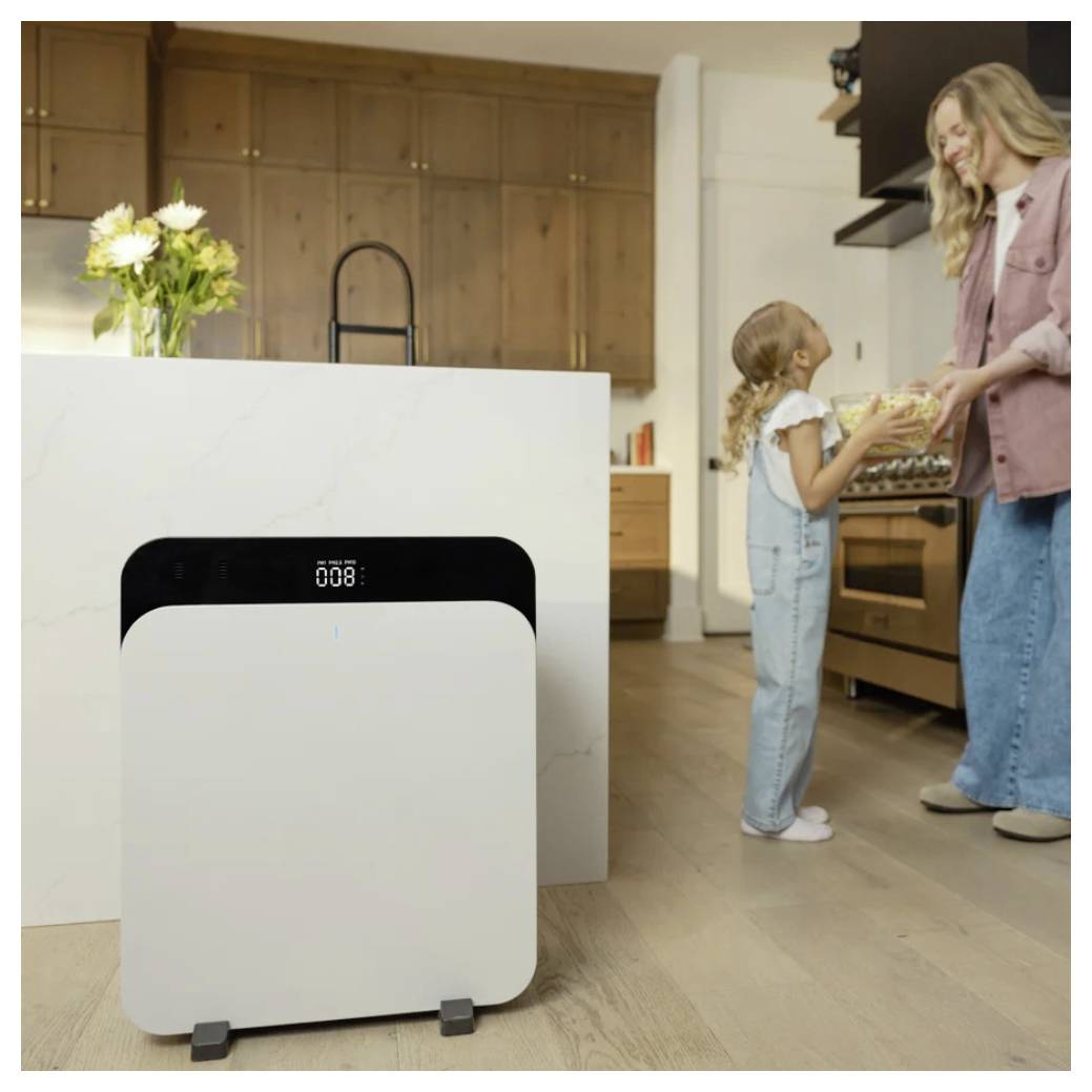 Ein Mädchen in Latzhosen und eine Frau in Freizeitkleidung stehen in einer Küche und lächeln einander an. Ein weißer Luftreiniger befindet sich im Vordergrund.